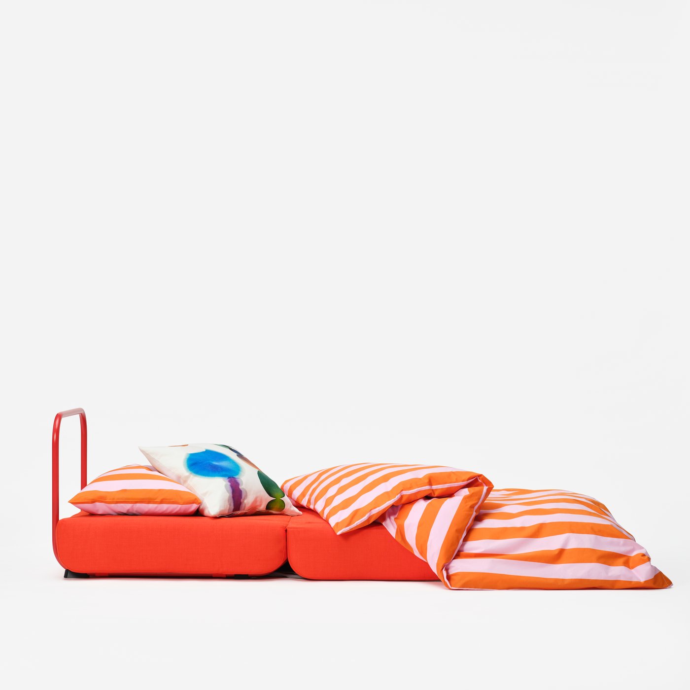 A red upholstered easy chair/bed, open with a striped duvet and coloured cushion on top, against a white backdrop.