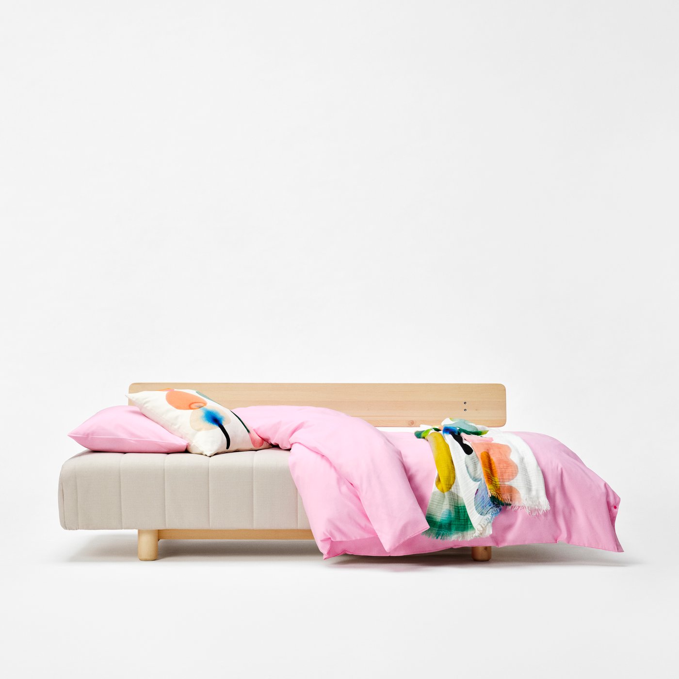 A beige sofa bed with a pink duvet and colourful textiles, against a white backdrop.