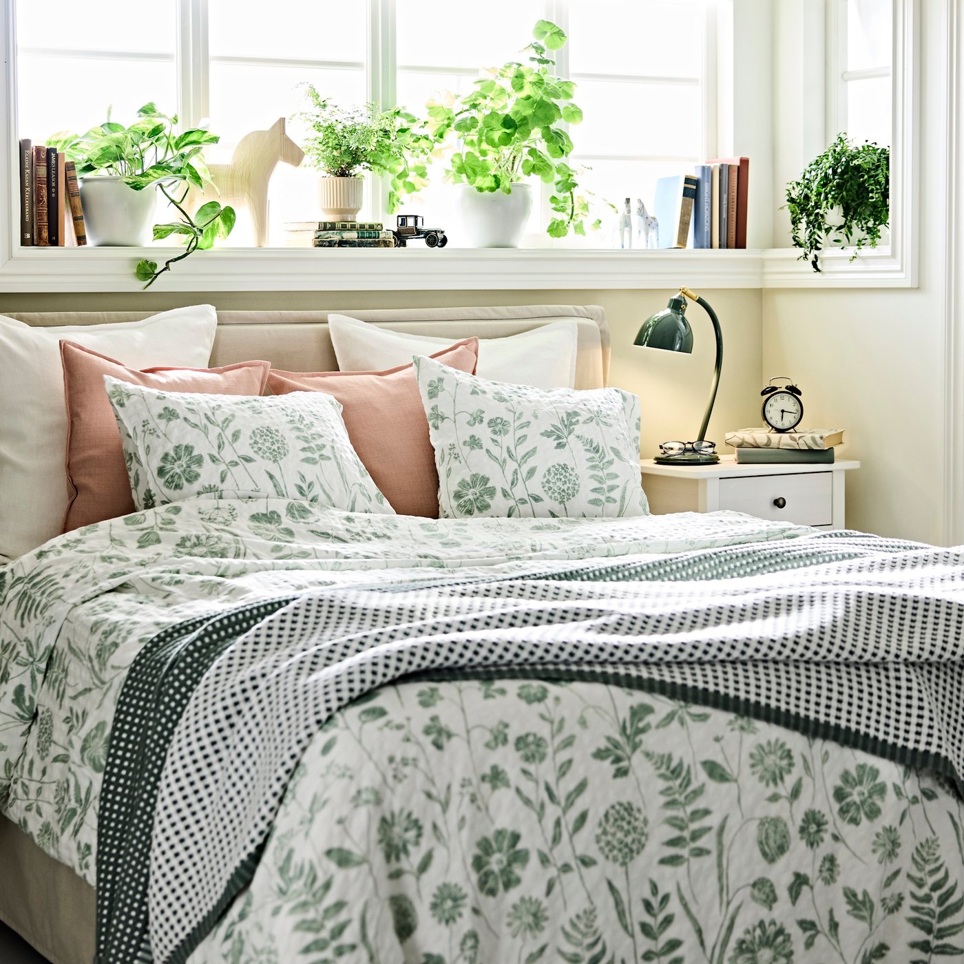 A bright, fresh bedroom with a bed dressed in botanical NÅLBJÖRNBÄR bed linen, up against a window ledge with many pot plants.