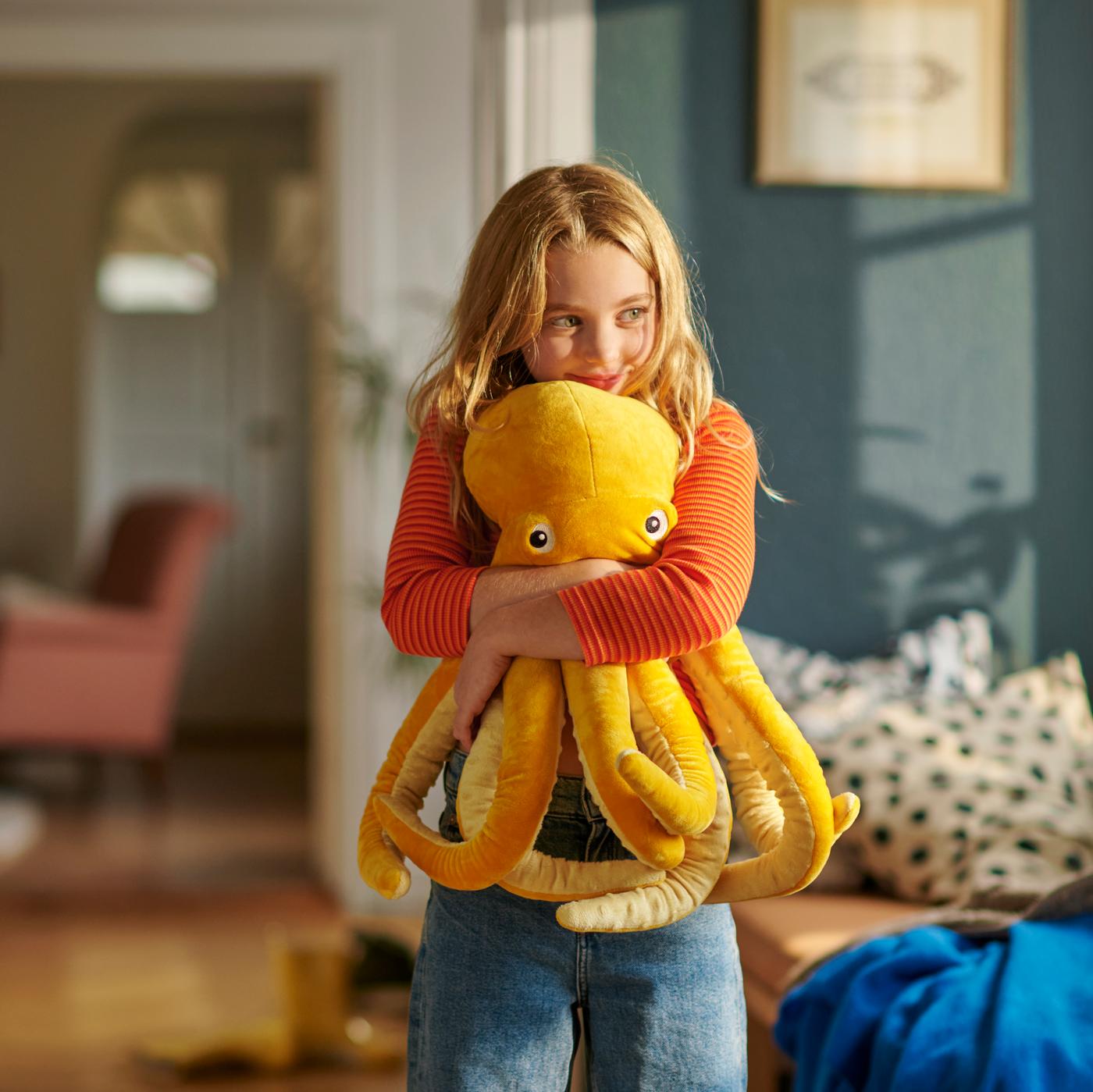 A child wearing an orange shirt and jeans, hugging the yellow BLÅVINGAD octopus soft toy.