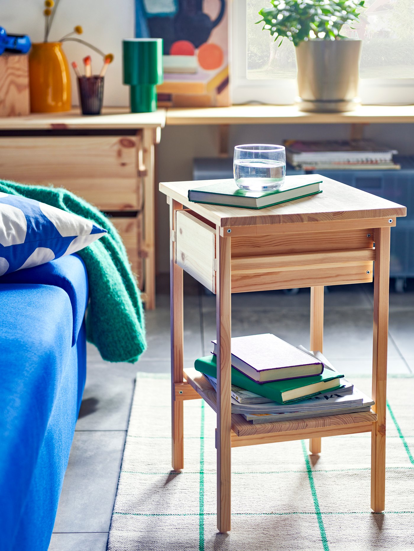 The KRONÖREN bedside table in pine, holding a stack of books on its shelf and a glass of water on top.