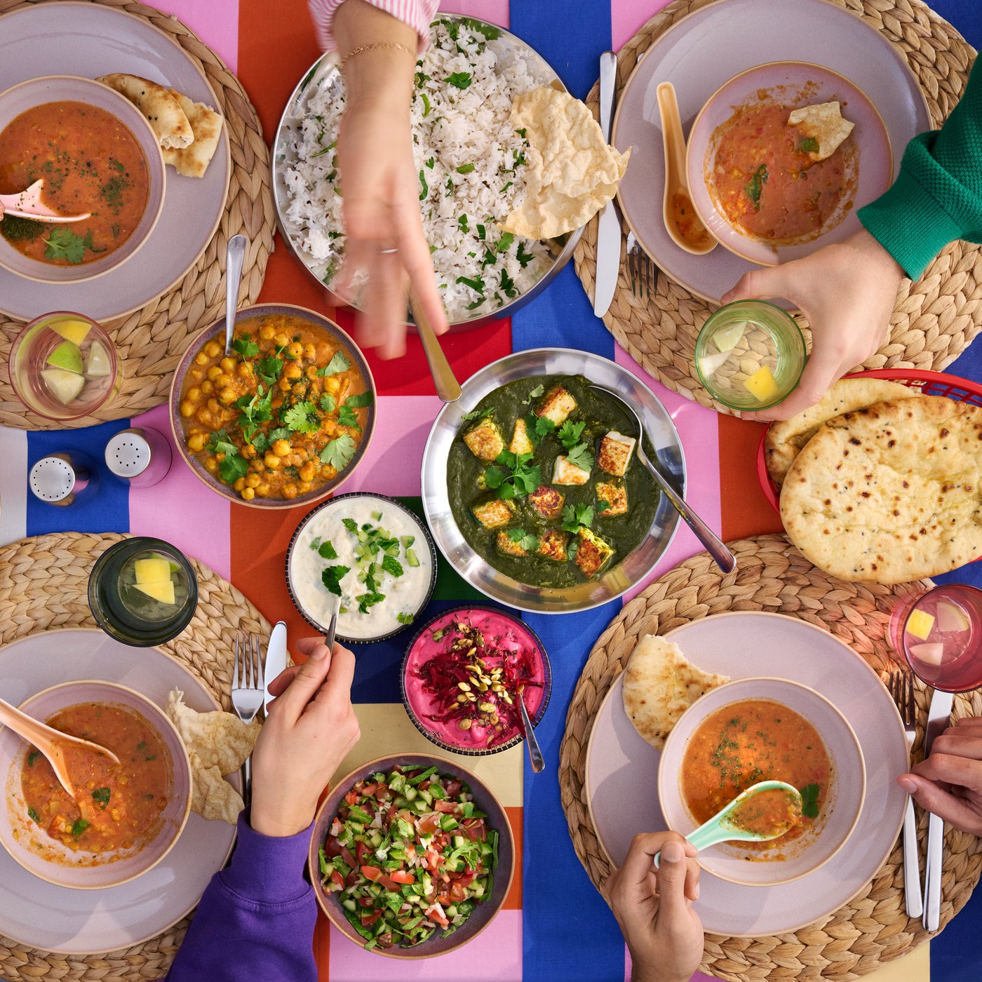 A colourful table setting with hands reaching for bowls of curries, rice, bread, and side dishes.