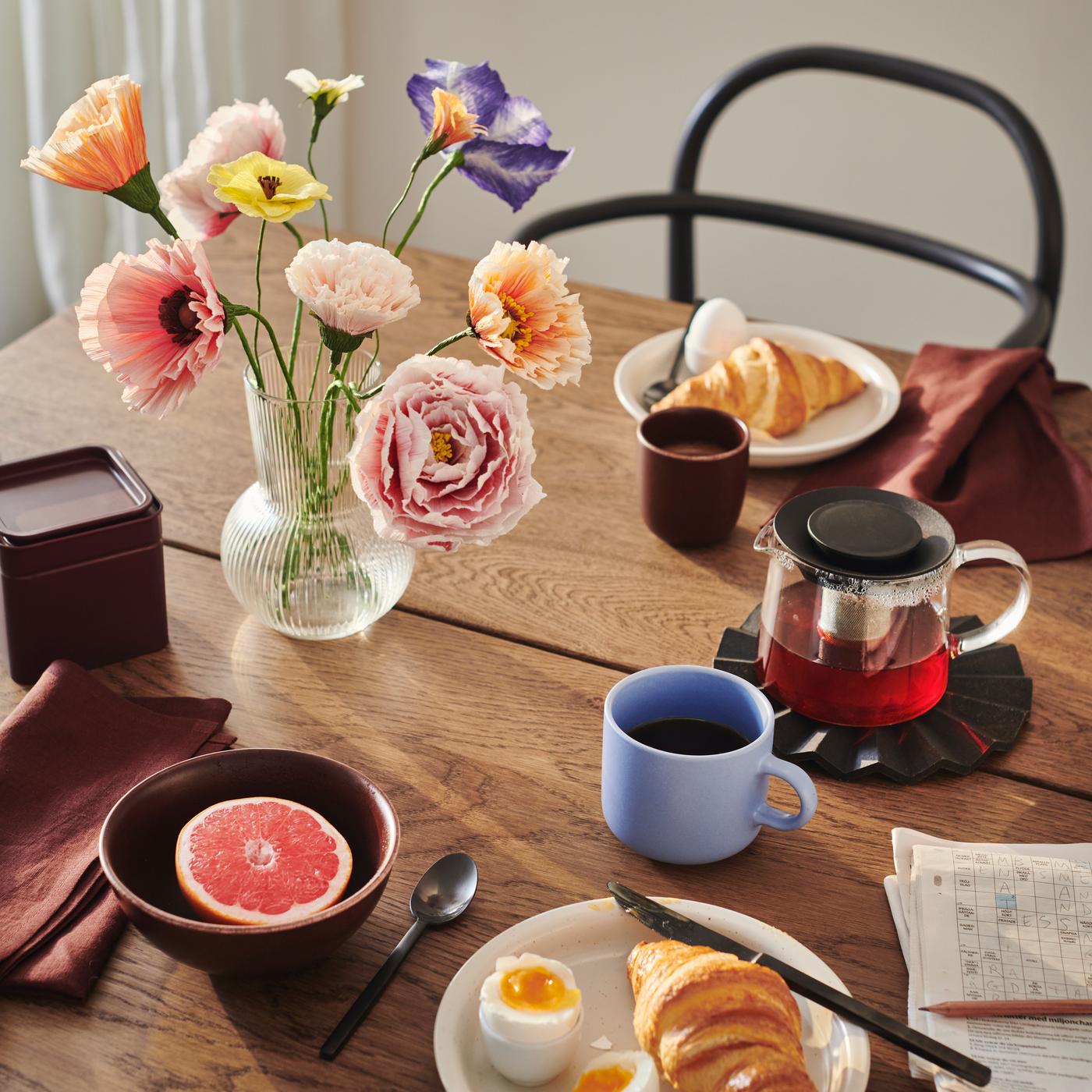 A wooden table set for breakfast, with a clear vase filled with colourful GULDSPETT artificial flowers.
