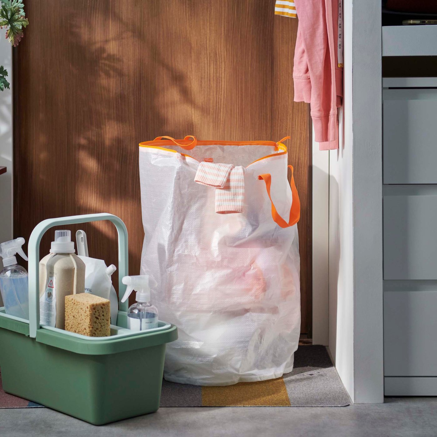A green bucket with handle full of cleaning tools beside a transparent laundry bag in a college dorm room.