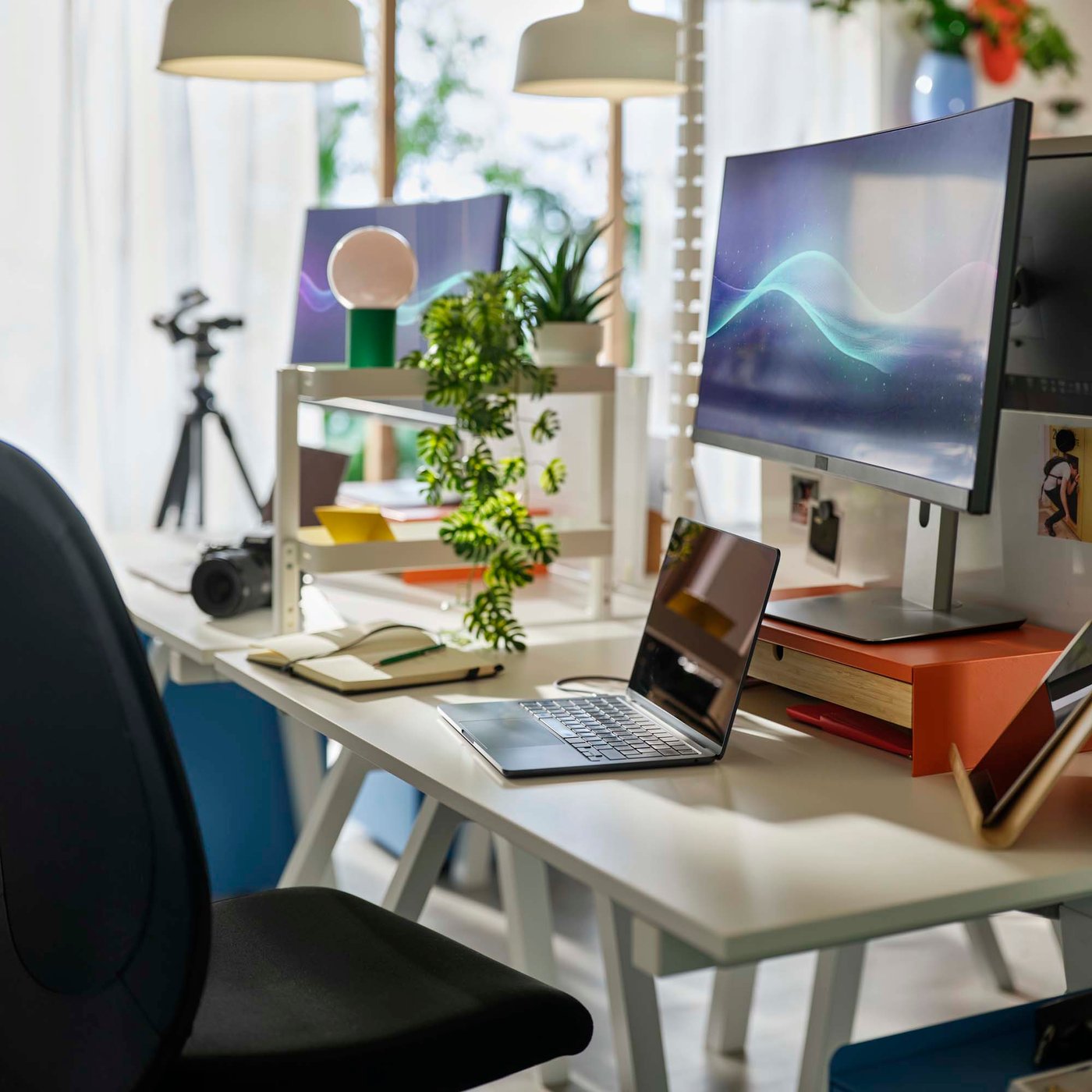 A creative desk area in a bright windowed room, with double monitors, a laptop and photo equipment in the background.