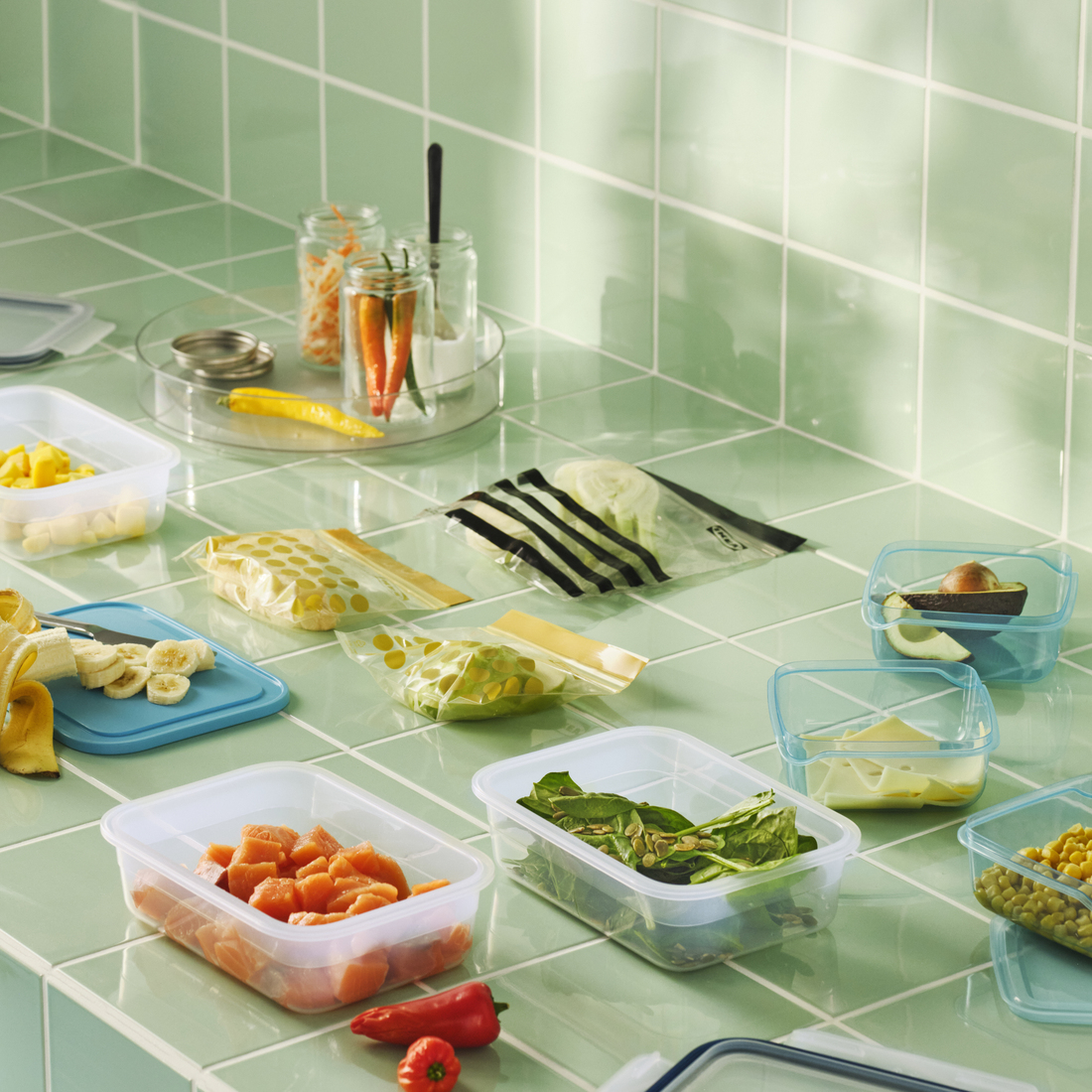Fruit and vegetables arranged in food containers and resealable bags on light green tiles. 