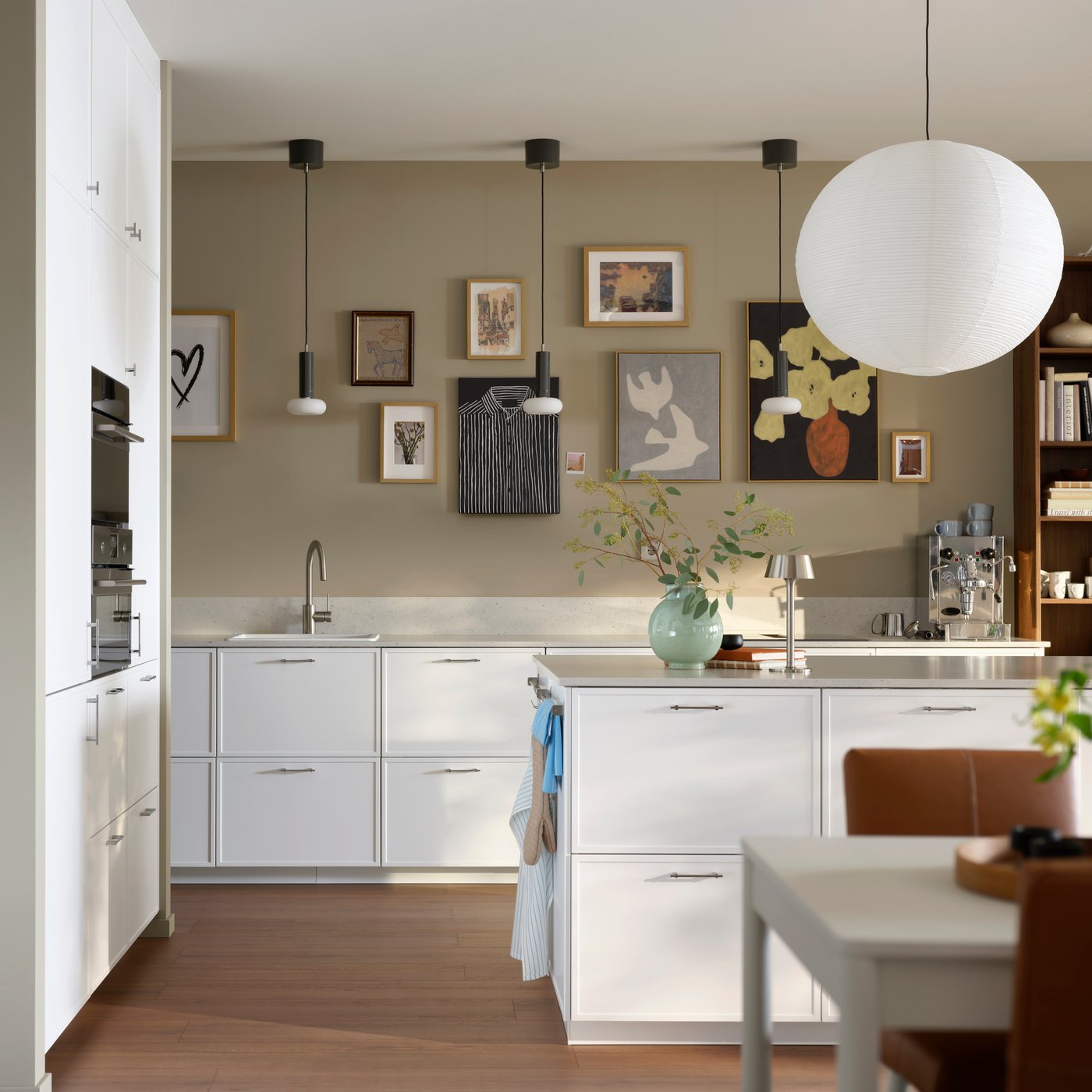 A modern kitchen with light grey walls filled with framed images and white minimal kitchen units. The sun casts shadows adding to the calm feel.