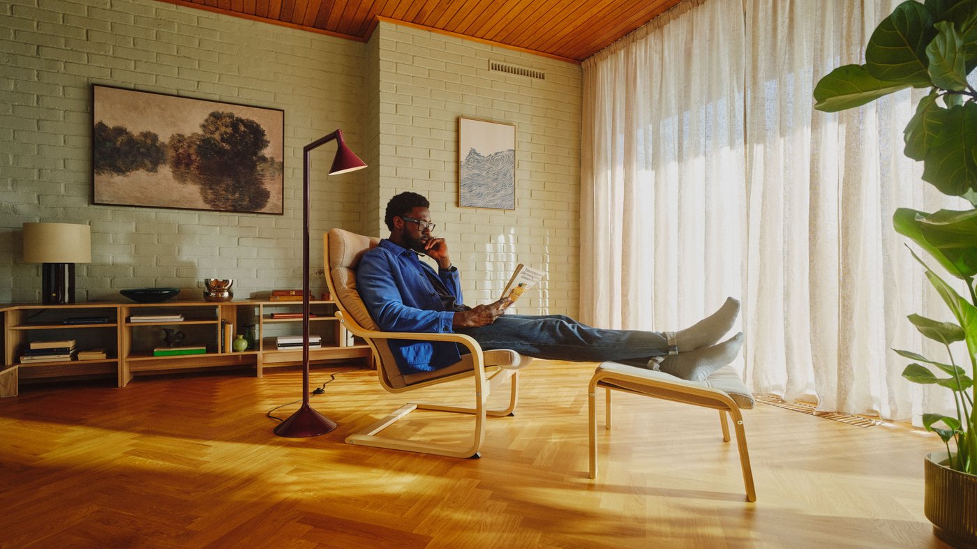 A person seated on a POÄNG armchair in the middle of a spacious living room with herringbone wooden floors, painted brick walls and sheer white curtains.