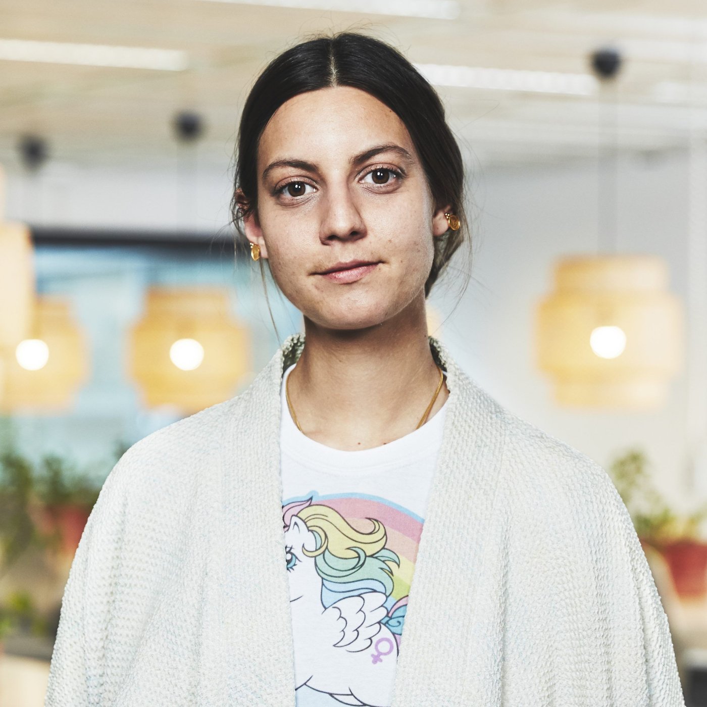 Portrait of Paulin Machado. The young woman is wearing a shirt and t-shirt. Illuminated lamps hang in the background.