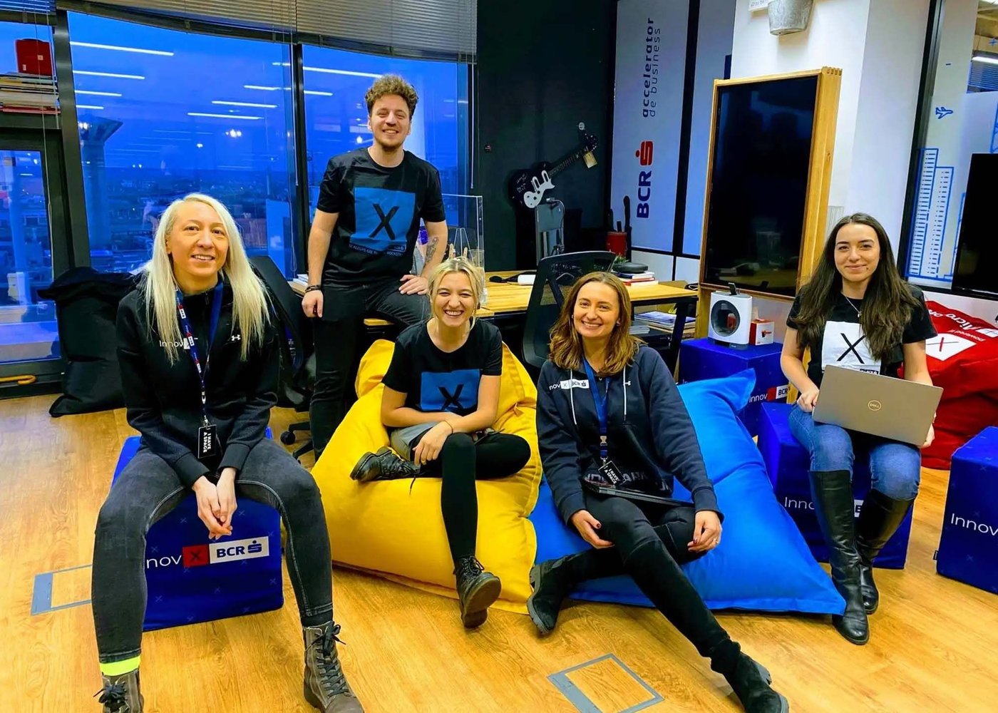 Two women sit on cubes, two people sit on beanbags and a man stands behind. They are all smiling at the camera. One woman is holding a laptop on her lap.