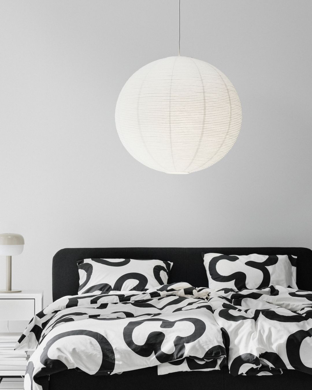 Behind the colour cutout of a GULLSUDARE pendant lamp hanging from above, a white room with a black and white bed appears.