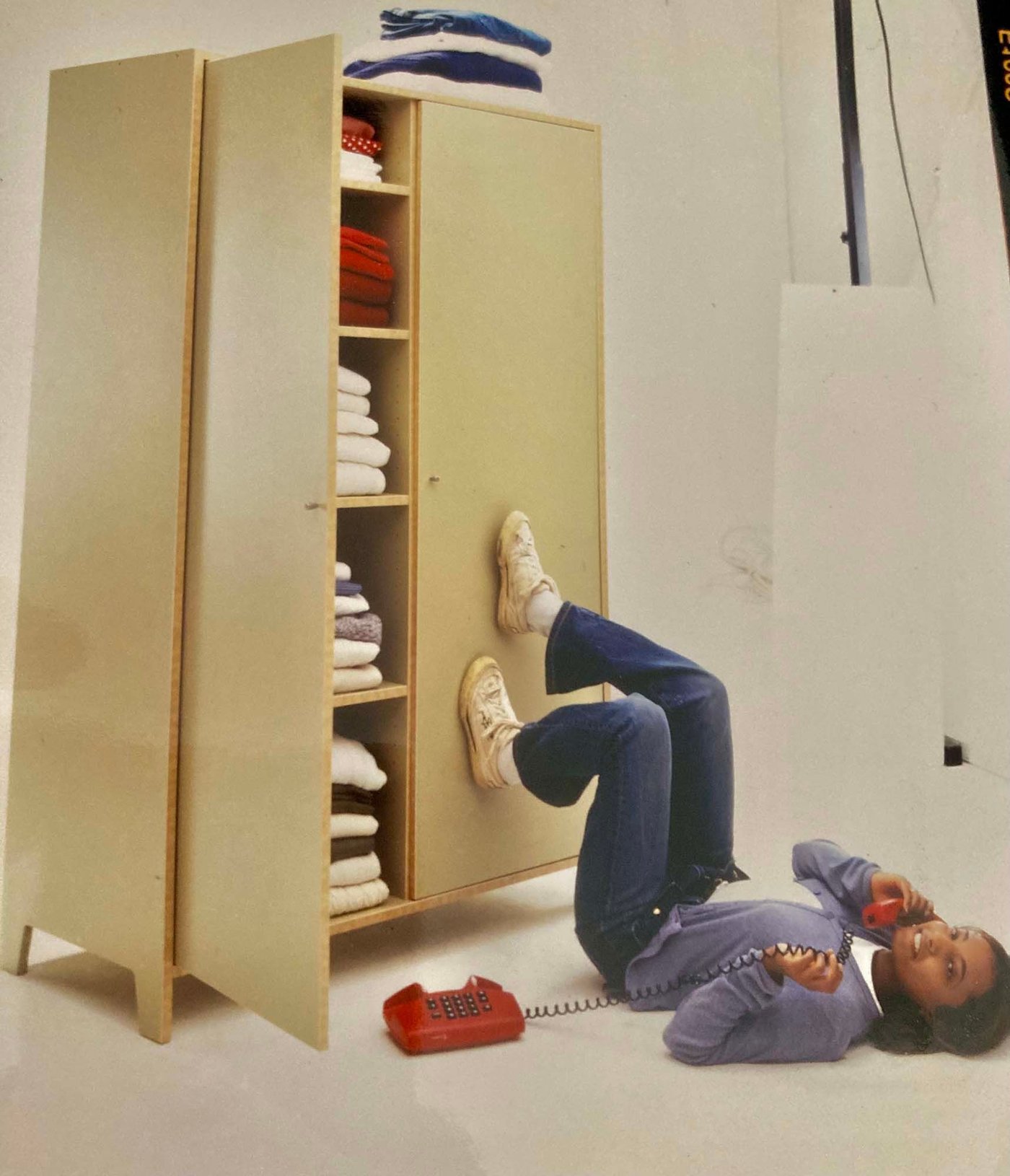 A vintage IKEA catalogue image of Rasika laying on the floor with her feet up against a cupboard, talking on a telephone. 