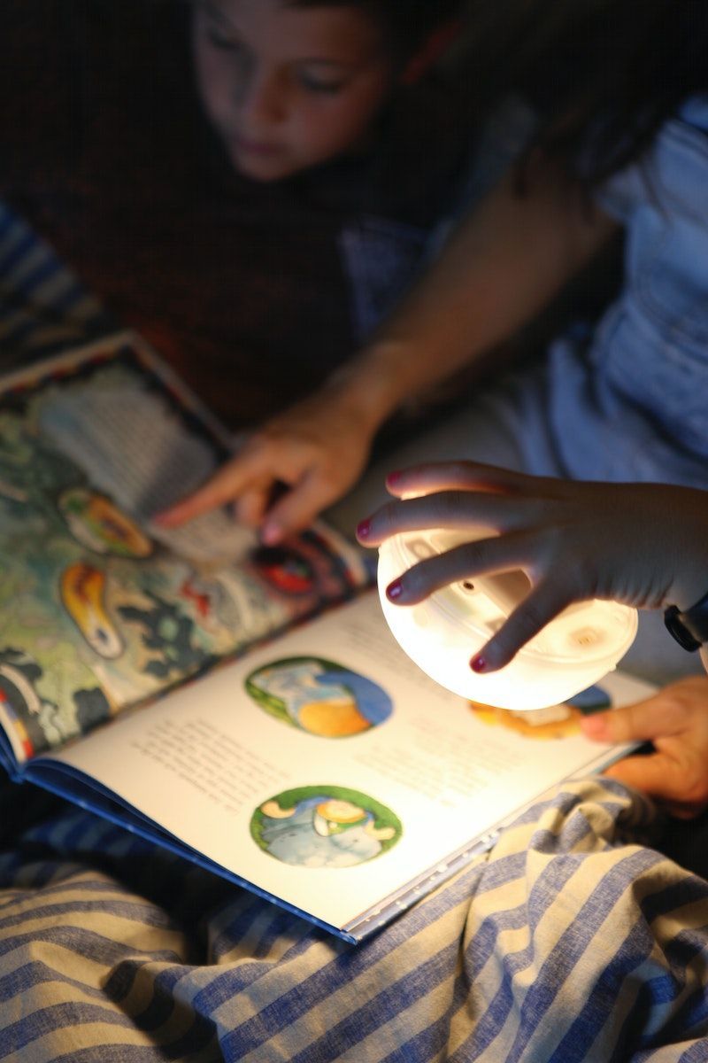 Children read a story book at night while holding the detached light source from the SAMMANLÄNKAD LED solar-powered table lamp.