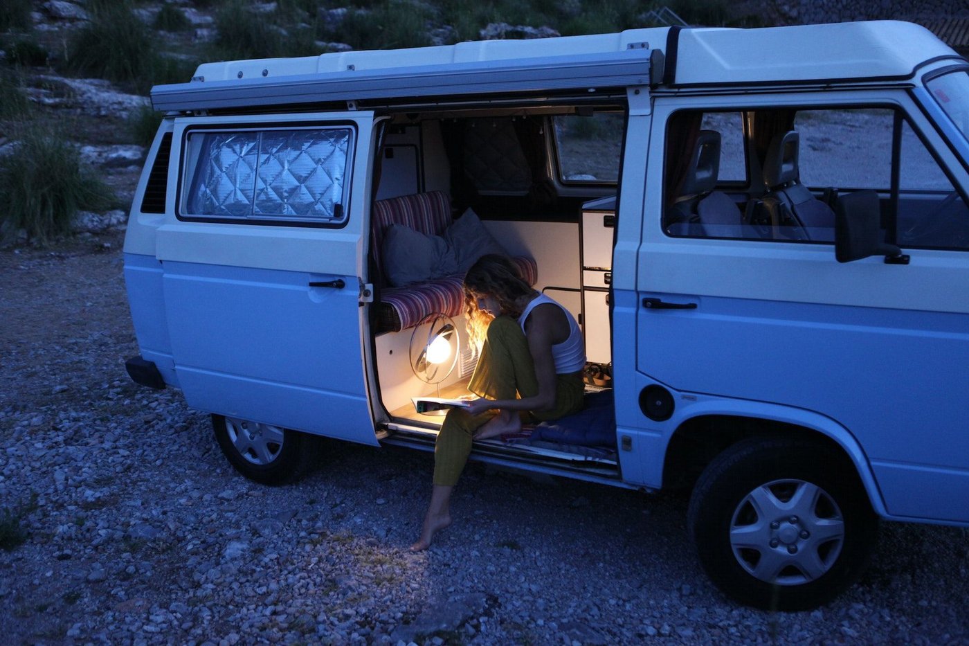 A person sits on the floor of an open-sided van at nightfall, illuminated by a SAMMANLÄNKAD lamp while reading.