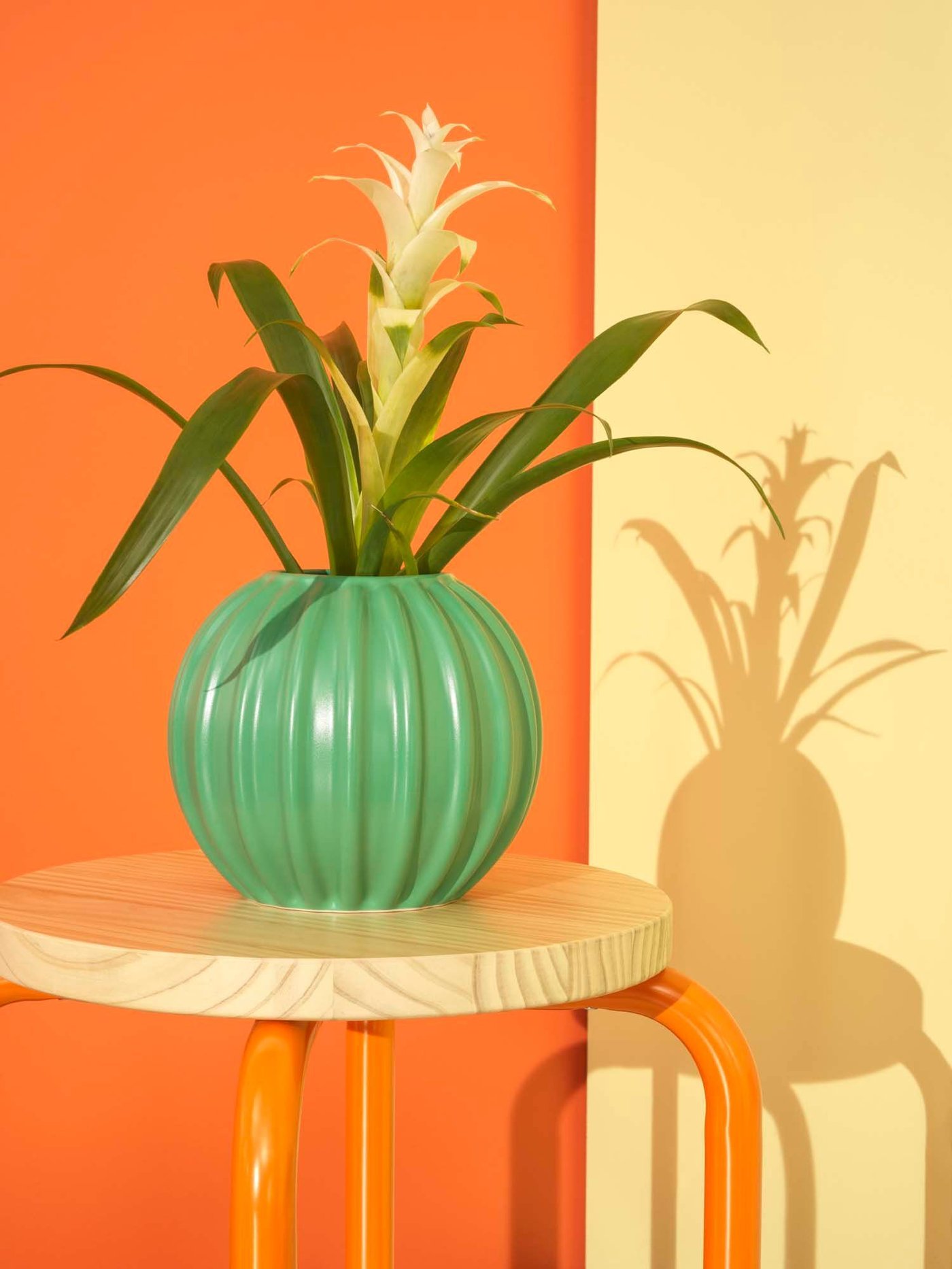 A green SKOGSTUNDRA vase filled with flowers sits on a stool, against a yellow and orange background.
