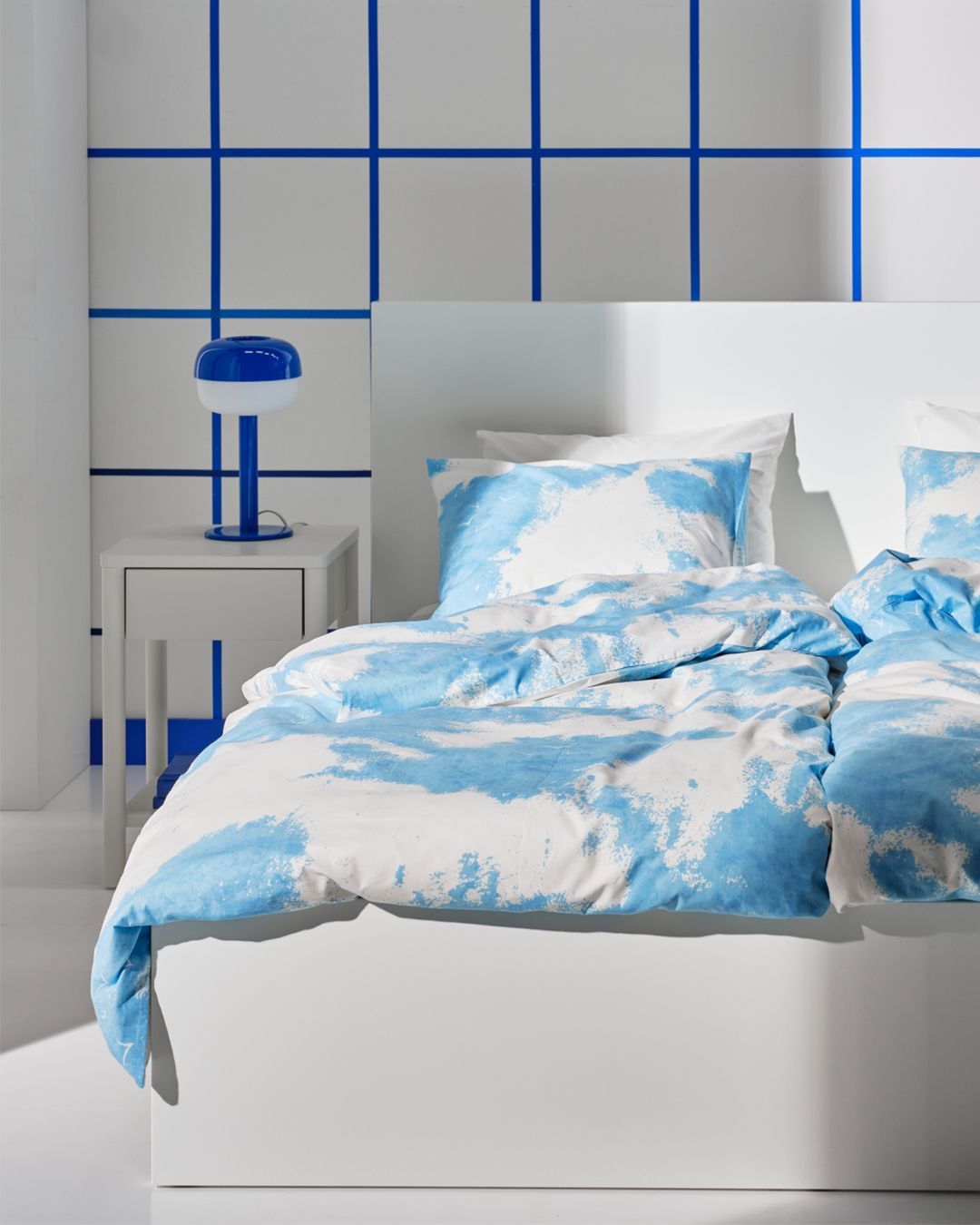The cutout of an  ÄNGSVITVINGE duvet and pillowcase with clouds transforms into a blue and white roomset in the background.
