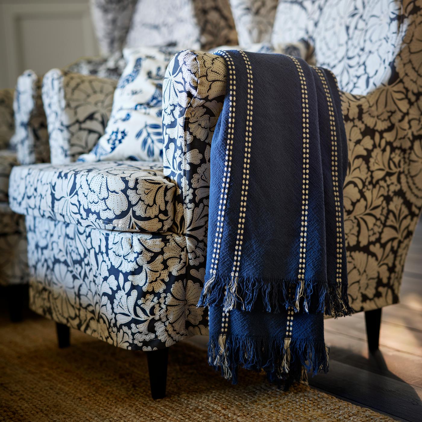 An elegant living room features a STRANDMON armchair with a floral pattern. Draped over the armrest is a dark blue fringed throw. The setting exudes a cozy and stylish vibe, inviting relaxation in the warmly lit space.
