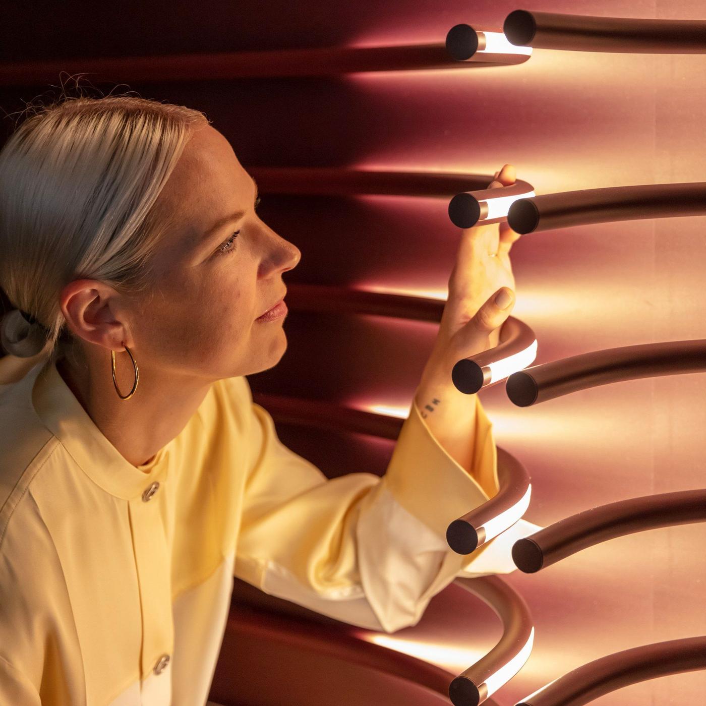 Designer Sabine Marcelis inspects curved lights from the VARMBLIXT collection, illuminated on a wall.