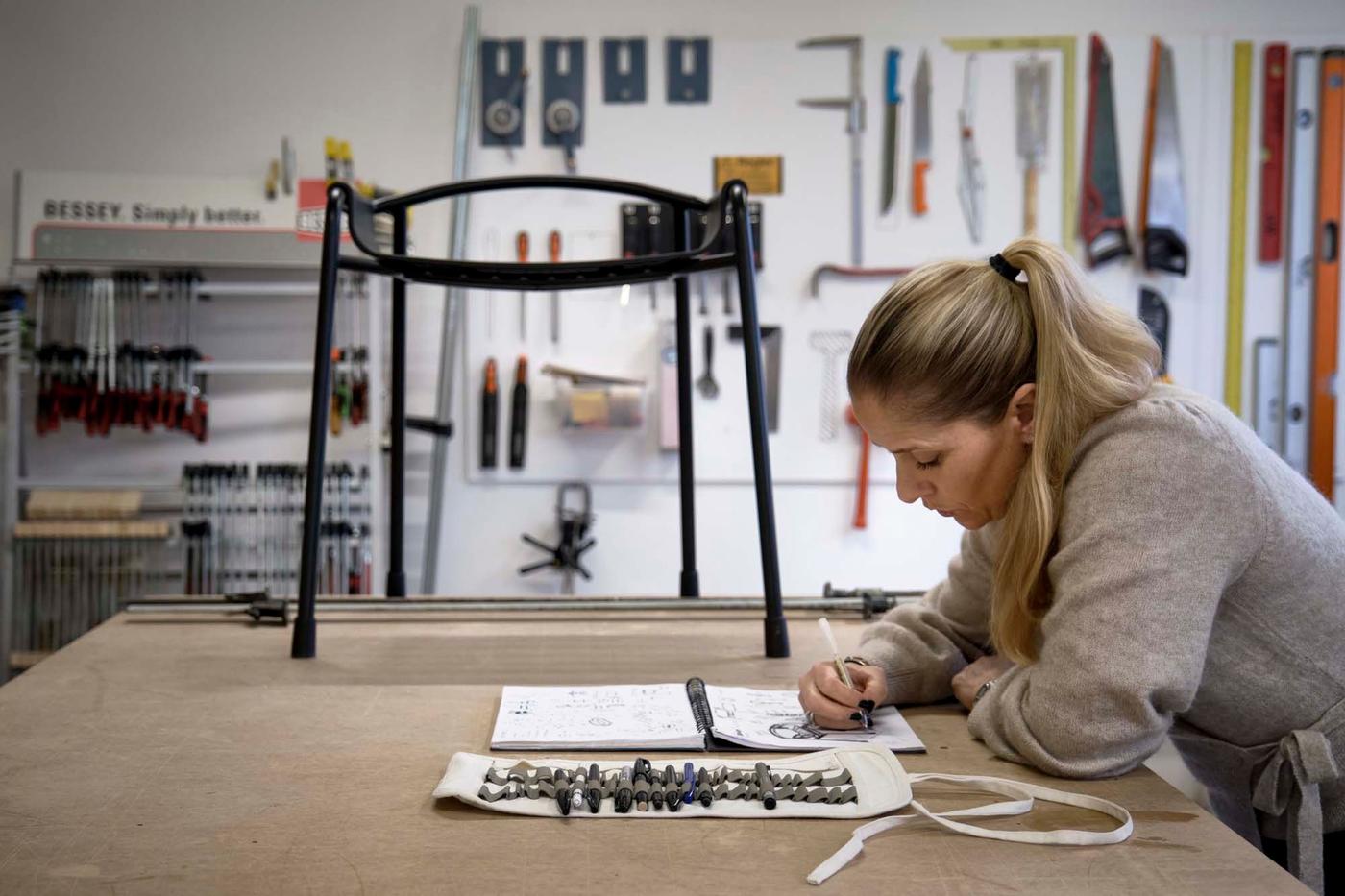 IKEA designer Sarah Fager sketches on a workbench with an anthracite BÄSINGEN shower chair in the background.