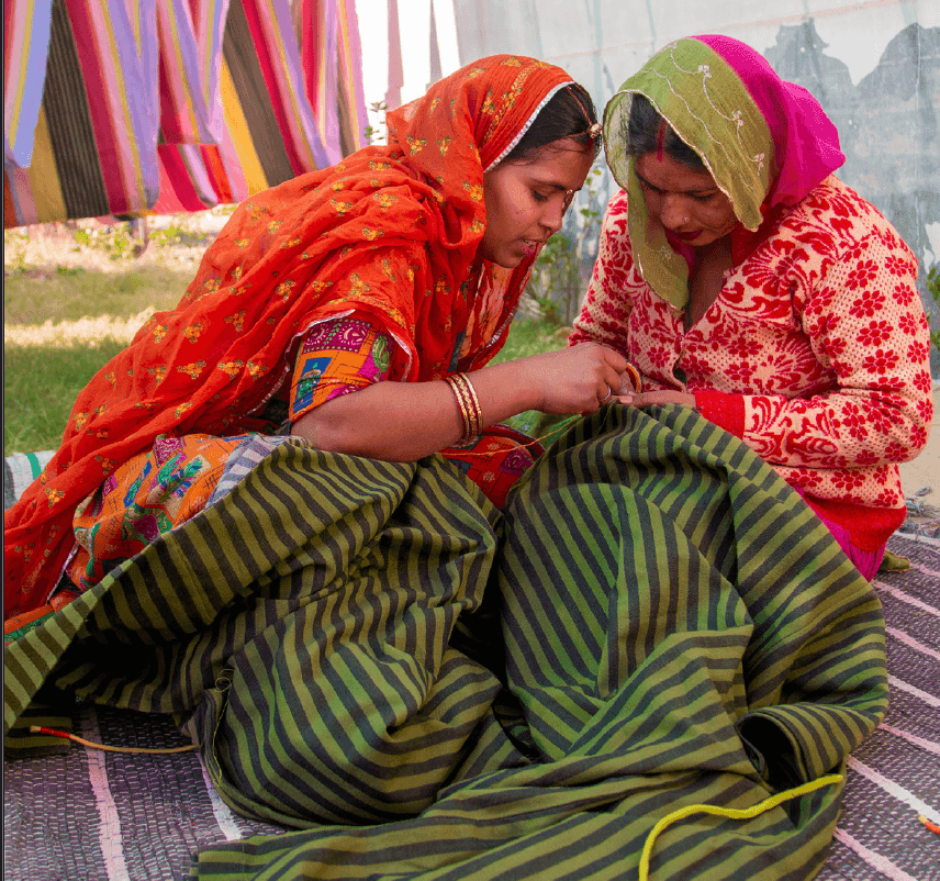 Two people sitting next to one another on the ground stitching a striped multicolour MÄVINN bath robe.