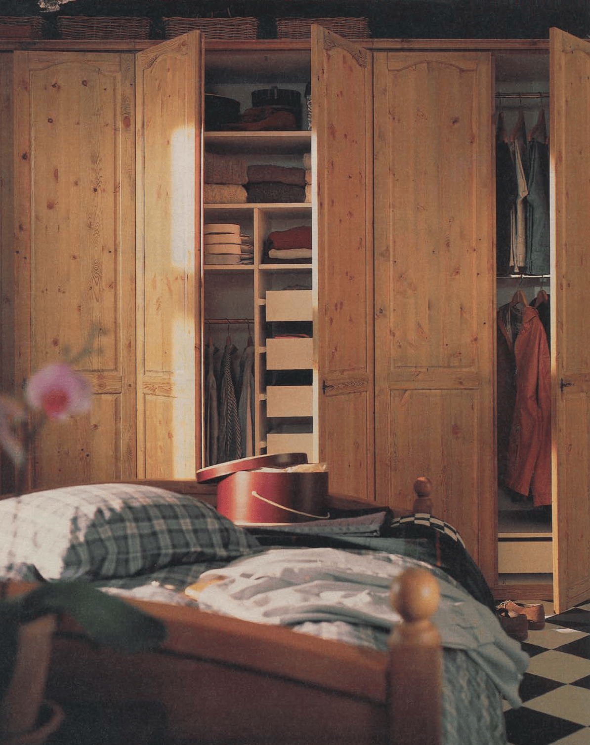 An open wooden wardrobe reveals neatly folded clothes on tidy shelves. In the foreground is a wooden bed.