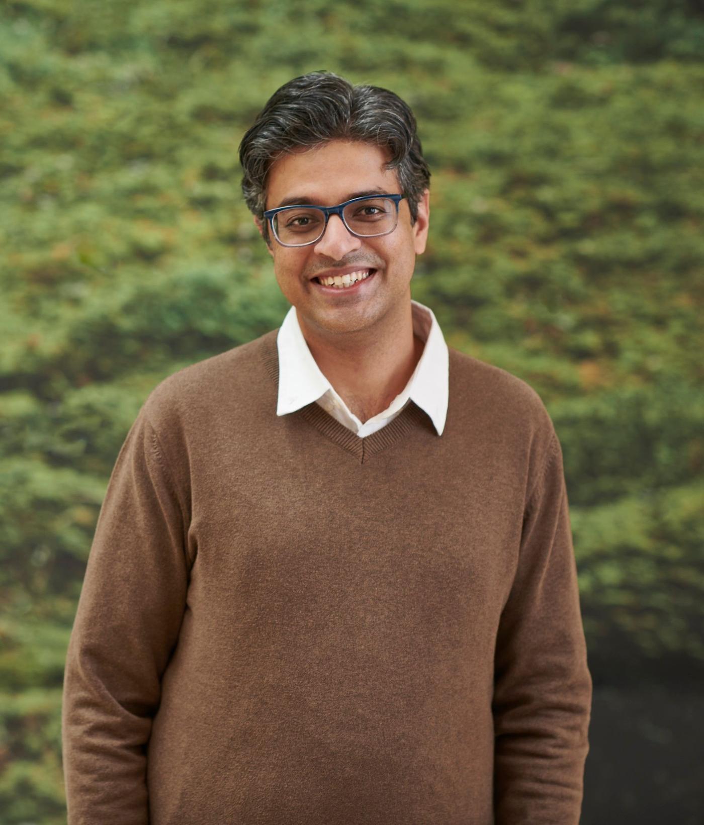 Portrait of Sriram Rajagopal, who is wearing a brown jumper and standing against a wall covered in green moss.