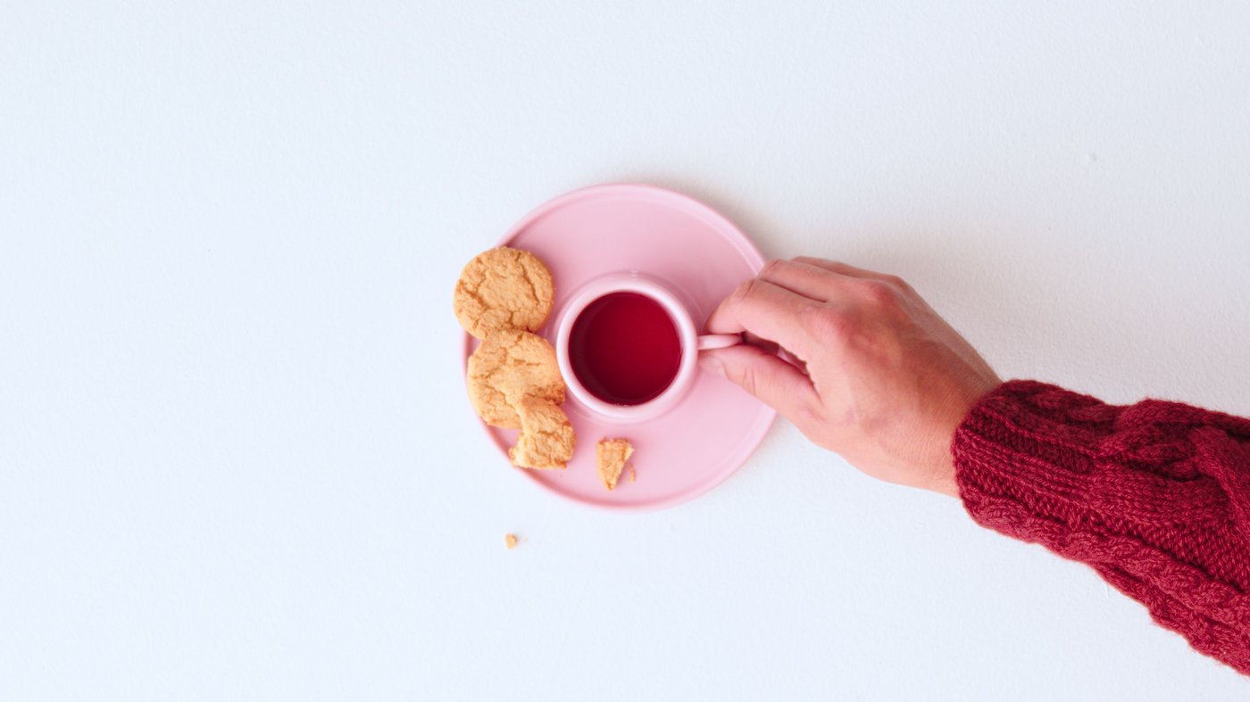A Pink cup seen from above with cookie crumbs around it, with a hand reaching for the handle.