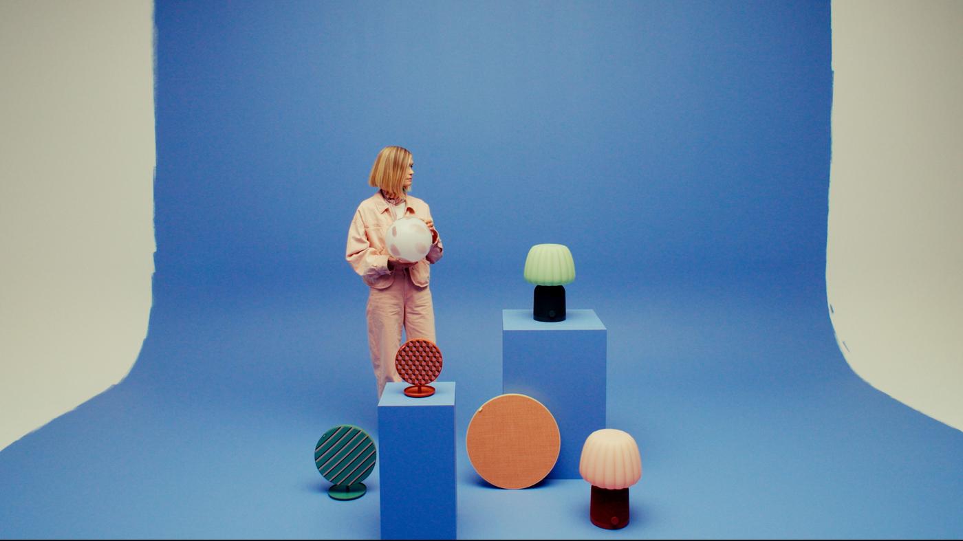 Tekla Severin stands among various speakers placed on pedestals, holding a spherical spotted lamp. The blue background is partially broken, revealing painted edges and unpainted white wall behind.