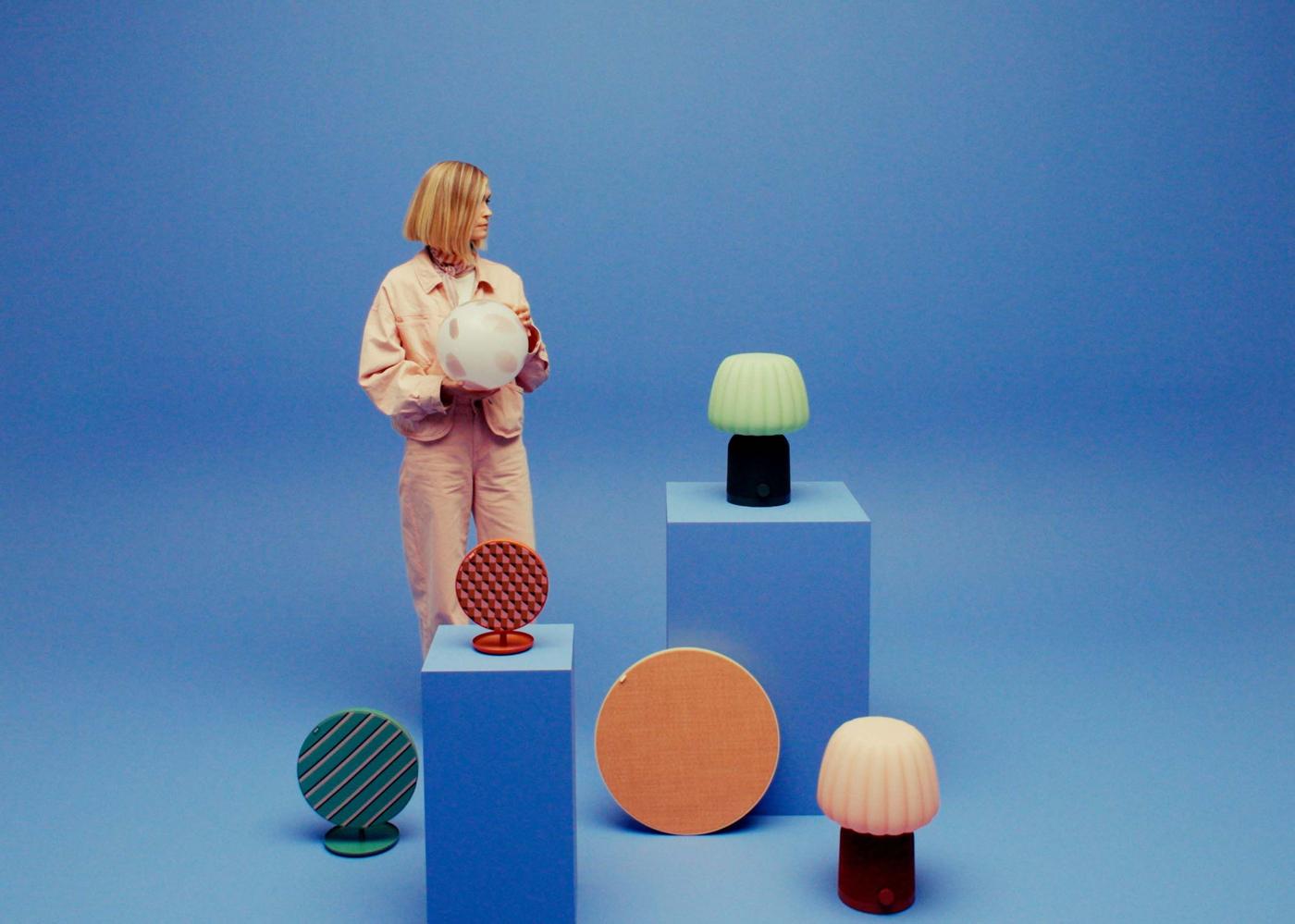 Designer Teklan Evelina Severin stands against a blue backdrop holding a lamp. She is surrounded by lamps and speakers on display.