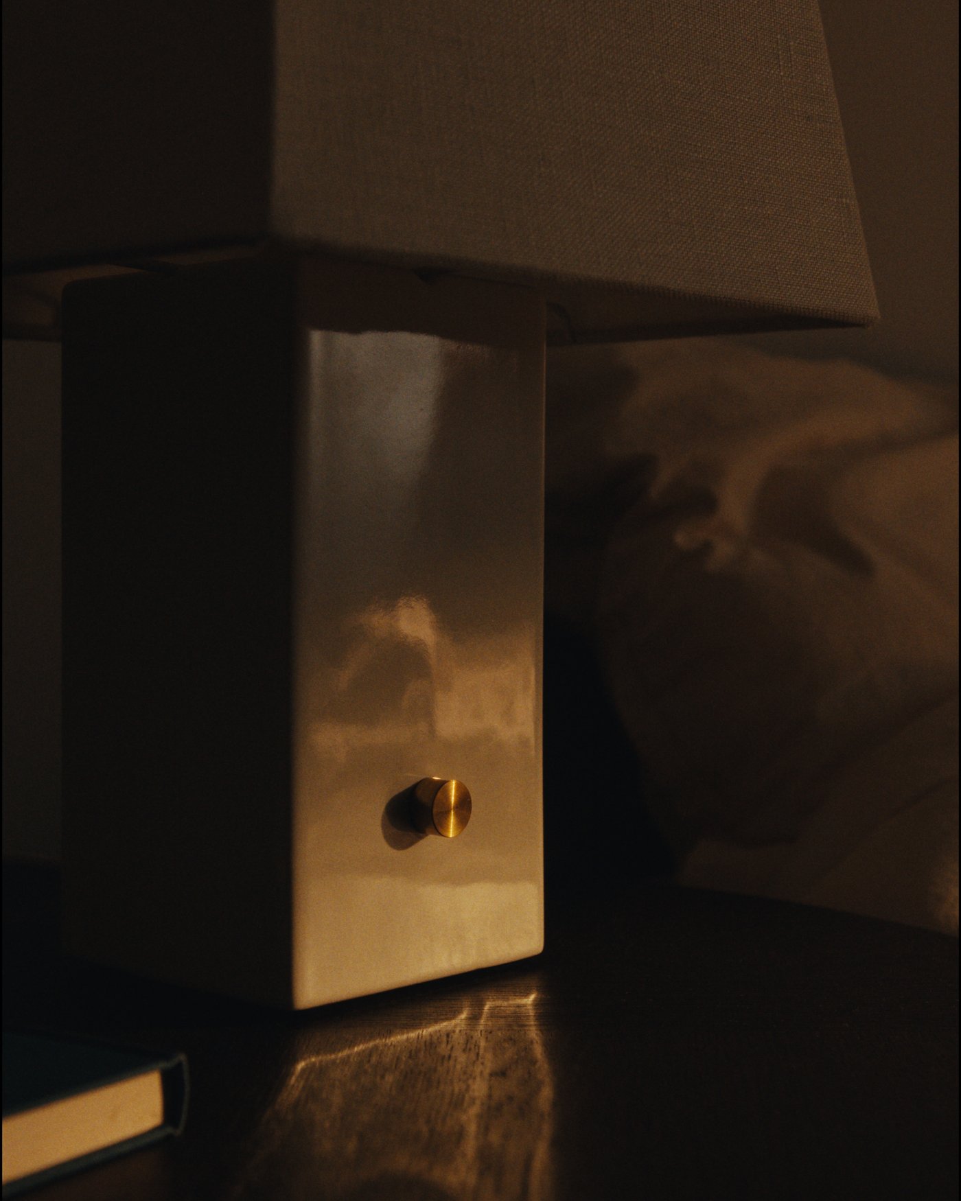 Close-up of a square ceramic lamp base with a linen shade and brass dimmer in low, warm light.