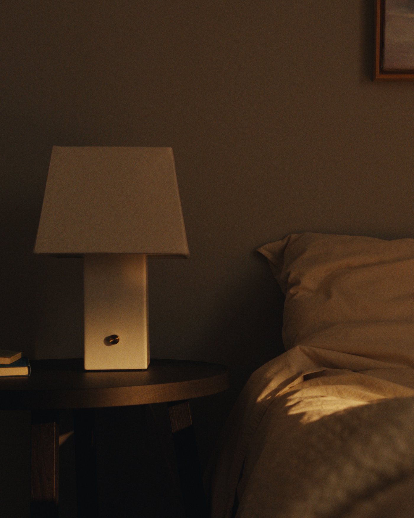 Square ceramic table lamp with linen shade on a bedside table next to a bed and stacked books.