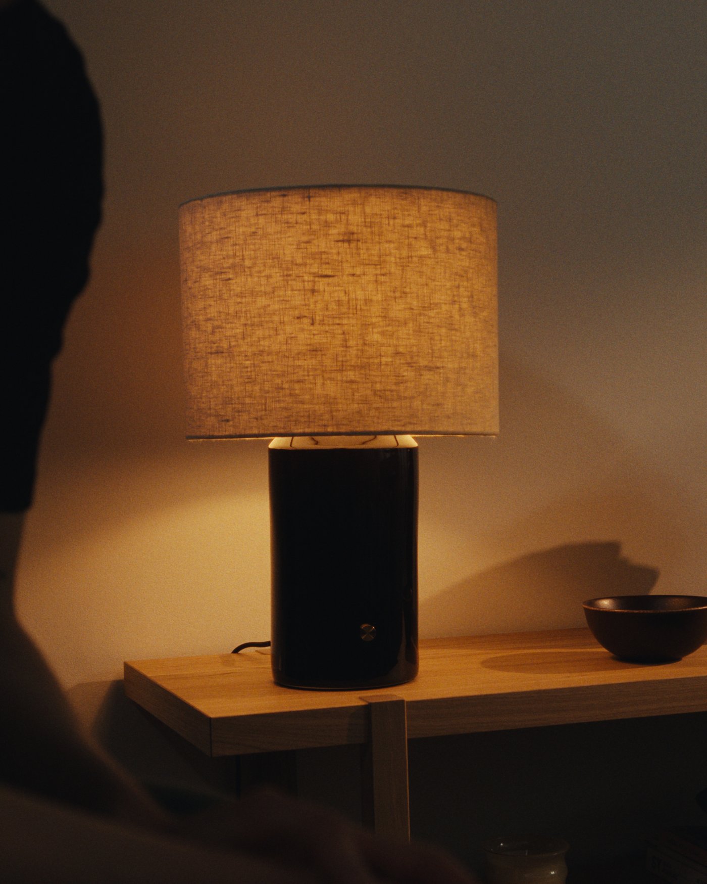Cylindrical table lamp with a dark ceramic base and linen shade on a wooden table, with a small bowl next to it and a person visible in the foreground.