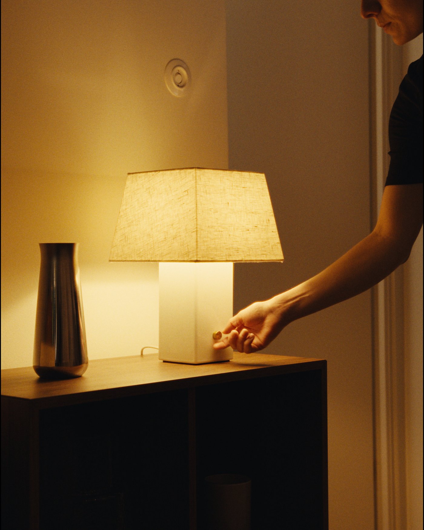 Hand adjusting the brass dimmer on a square ceramic table lamp with a linen shade.