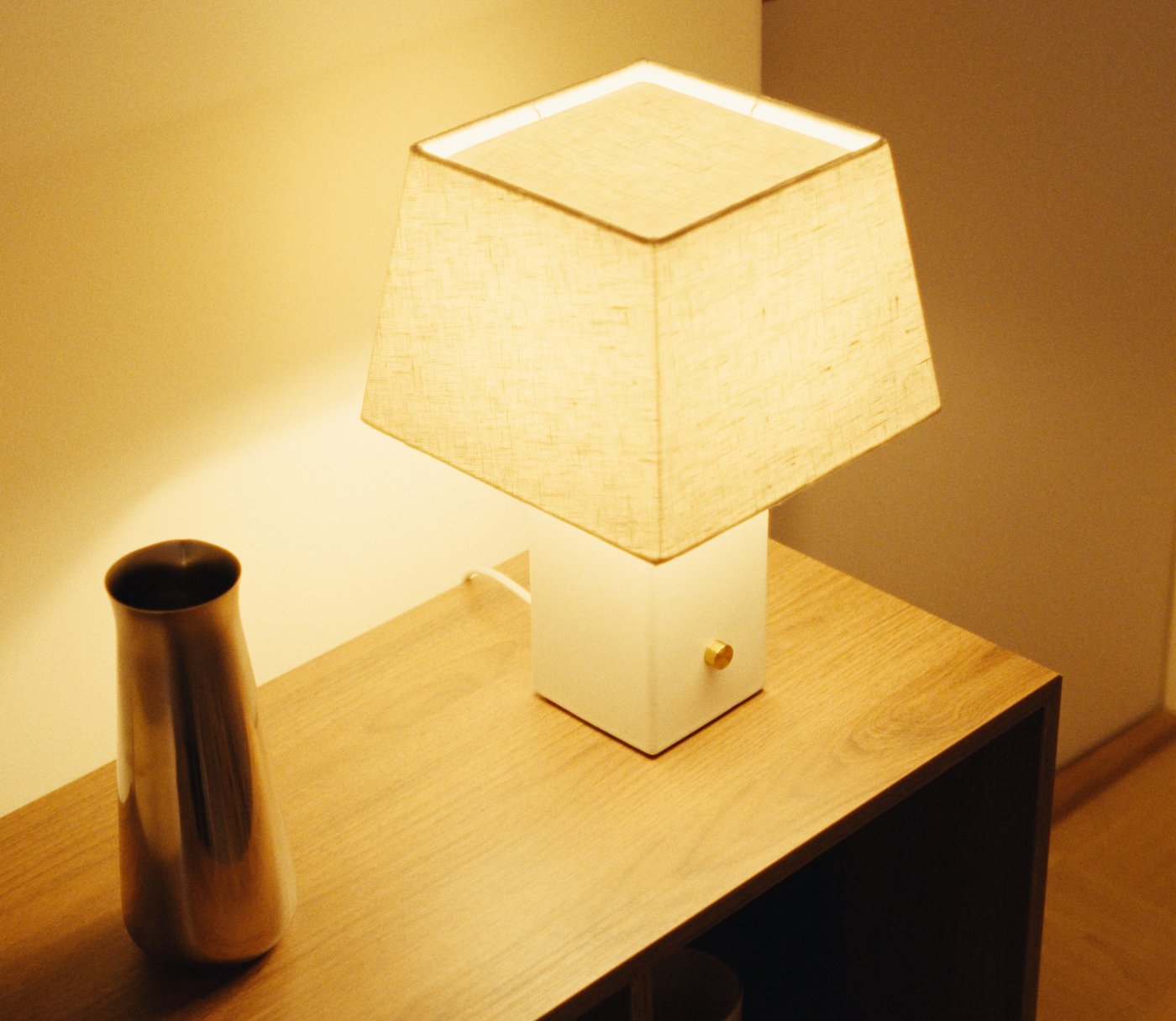 Square fabric-shade table lamp on a wooden console with a metal vase beside it.