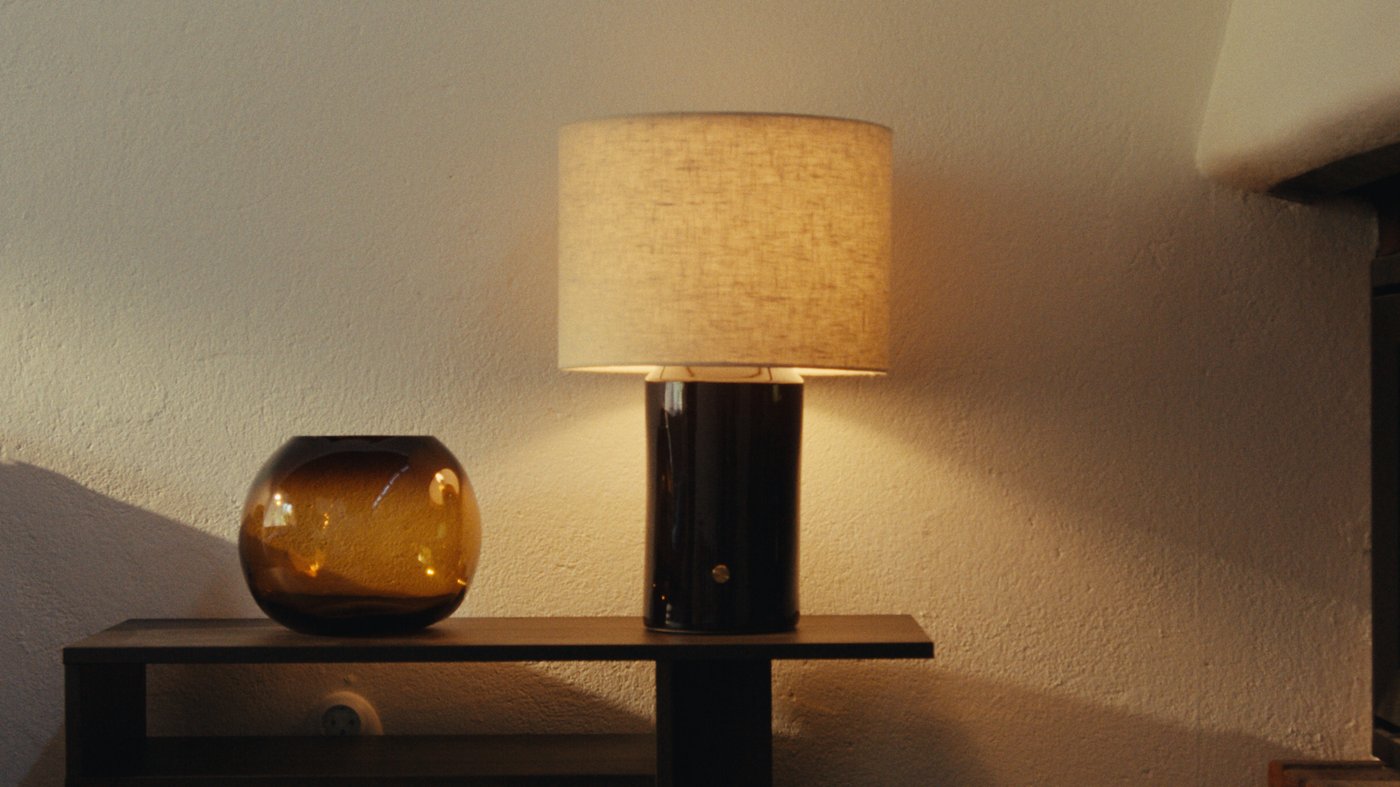 Table lamp with a beige fabric shade and dark cylindrical base on a shelf, next to an amber glass vase against a textured wall.