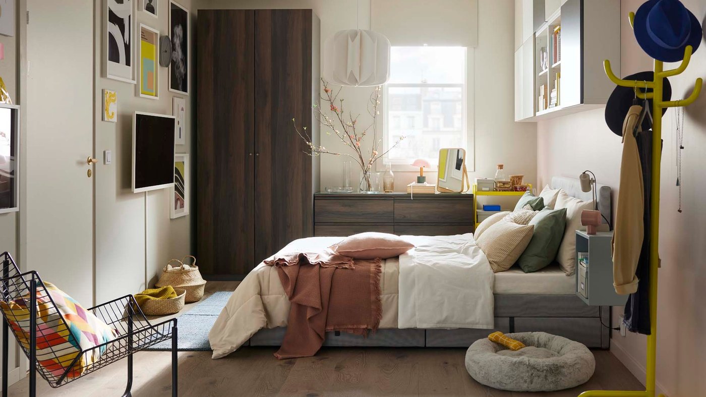 Slider image one: a wide angle view of a bedroom style in pale neutrals with dark wood cabinets, and accents of bright yellow and soft pinks.