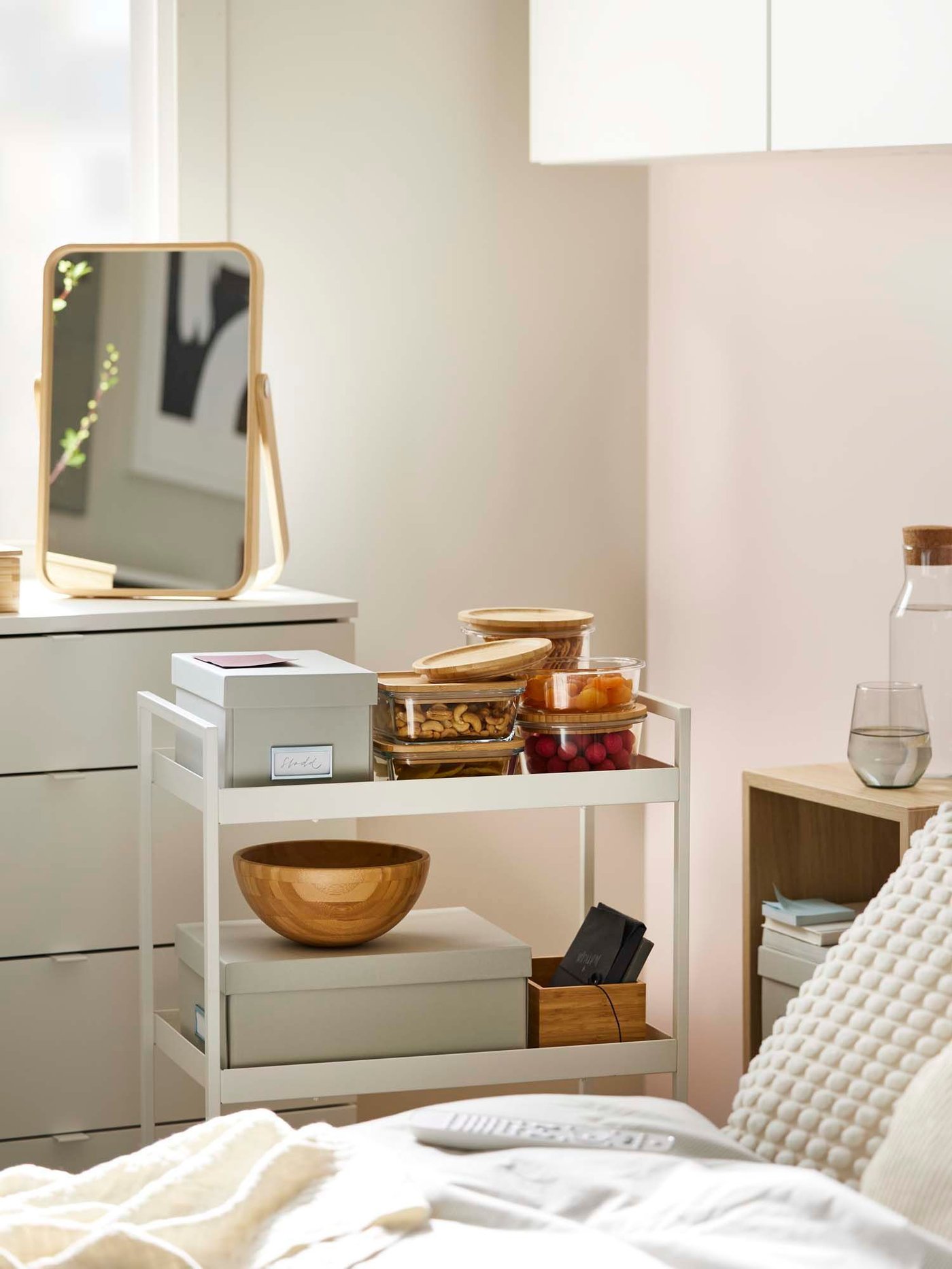 Slider image two: a white NISSAFORS trolley pulled up alongside a bed, style with storage boxes, glass food containers filled with snacks and wooden accessories.