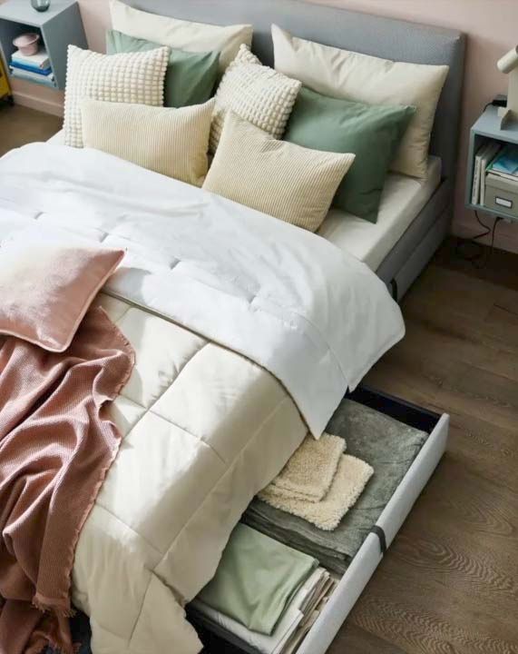 A high-angle view of a double bed in soft pastels and neutrals, with underbed storage drawers open revealing bedlinen.