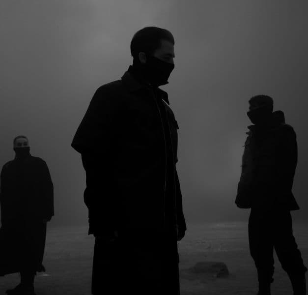 In a dimly lit grey space, the silhouettes of three people standing.