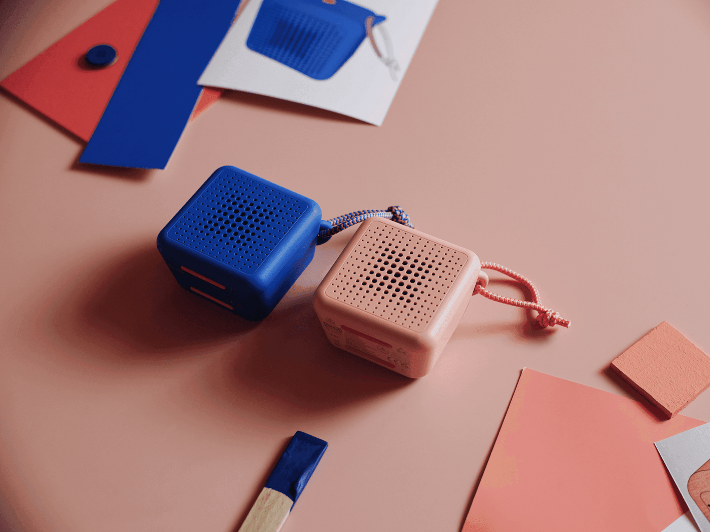 Two VAPPEBY portable bluetooth speakers, one in blue and the other in pink, are lying down on a peach coloured desk, with blue and peach paper stripes lying next to them.