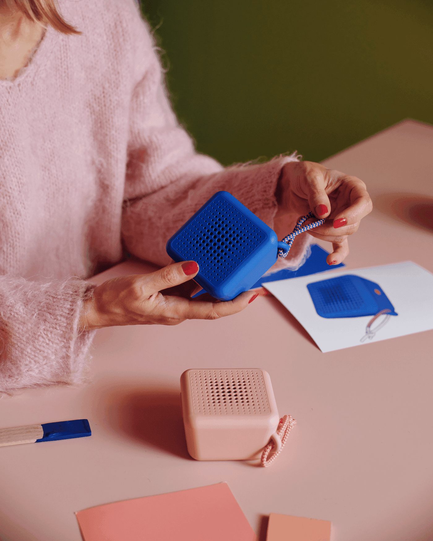Shot from above, a woman in pink is seen to handle and turn a blue portable speaker over a peach desk. Pink bluetooth speaker lies with its back down facing upwards. Next to it, is a blue sketch of the blue speaker.
