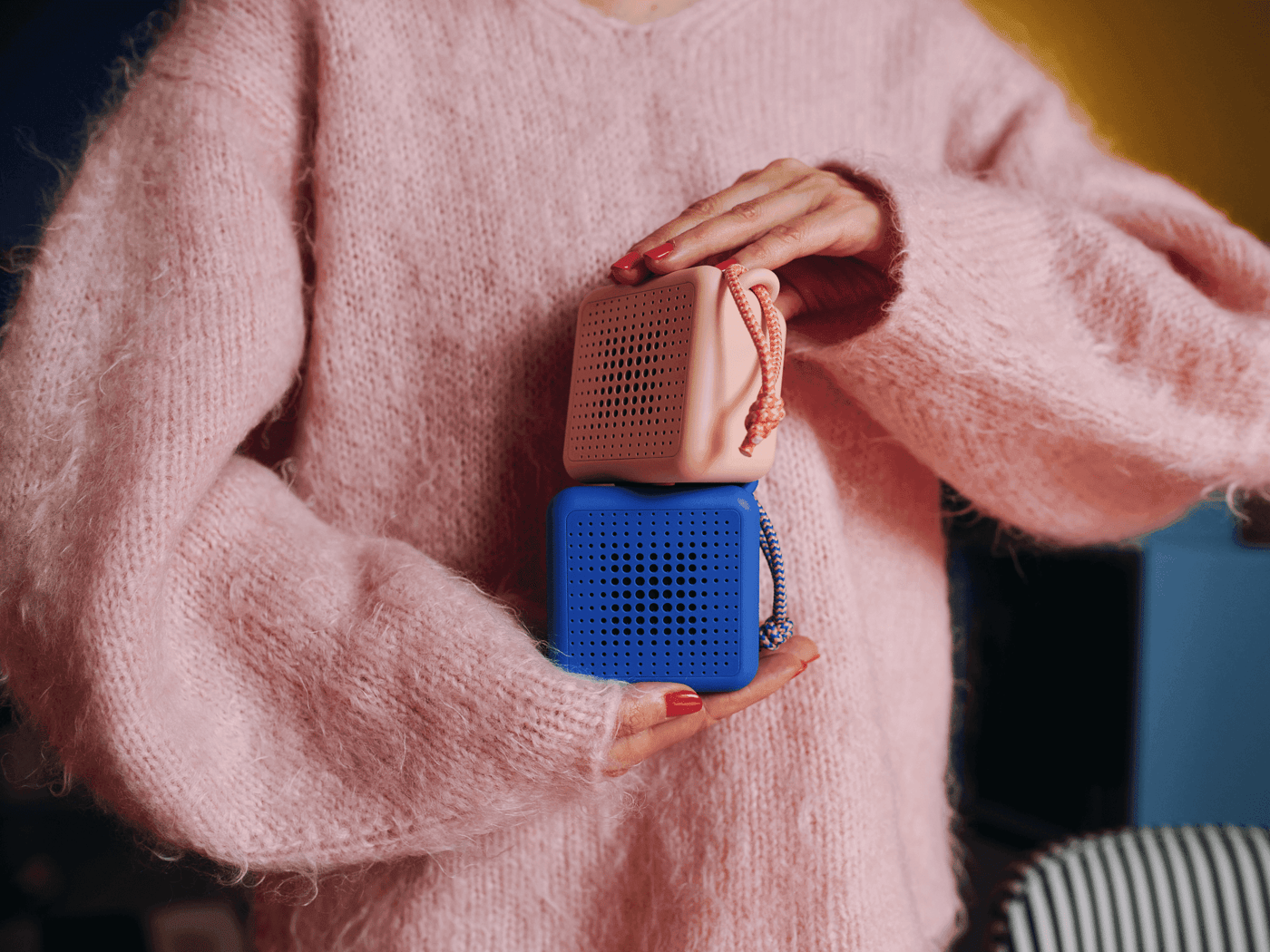 A person dressed in a pink-coloured jersey is holding two portable bluetooth speakers, one in blue and the other in pink, on top of eachother, holding them between upper left hand and lower right hand.