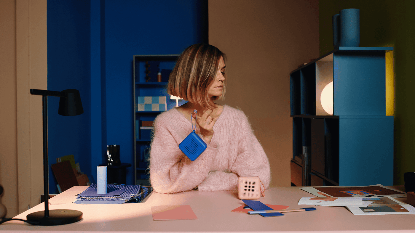 Multidisciplinary designer and colourist Teklan sits at desk, in pink and turned away, holding a blue portable speaker in her right hand, with a pink speaker to her left. 