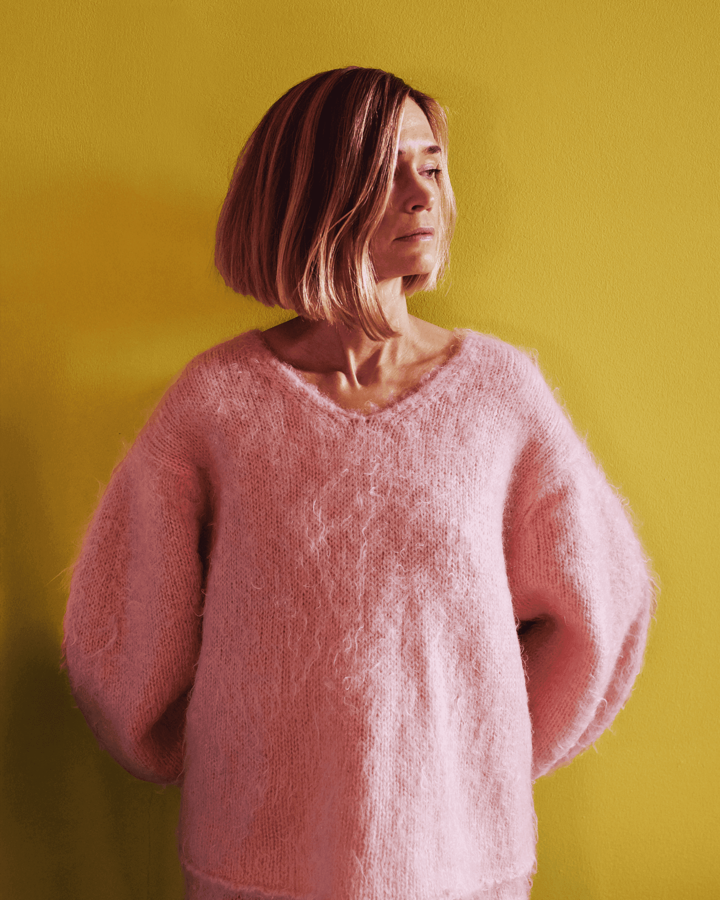 Tekla Evelina Severin standing with a pink outfit against a mustard yellow wall, looking towards the side.