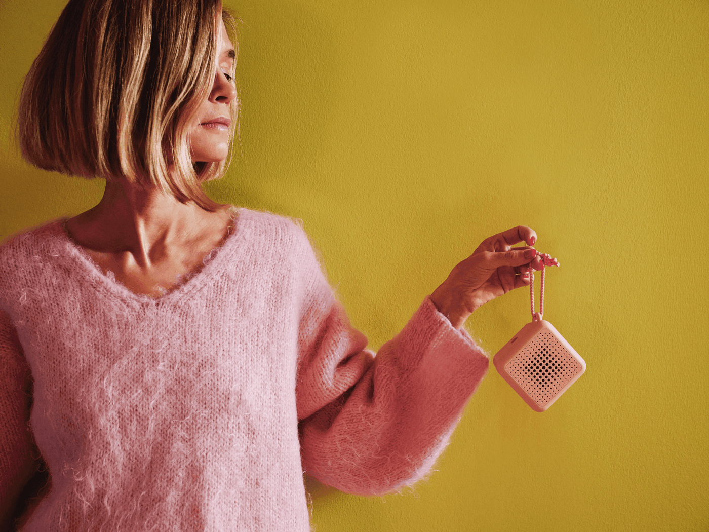 Tekla Evelina Severin is dressed in pink standing against a yellow wall holding up a small pink portable VAPPEBY speaker, holding it's leash.