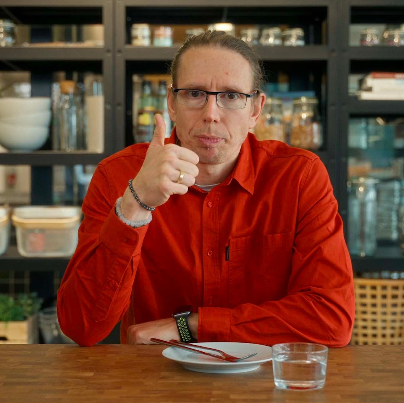 József sits at a table with an empty plate in front of him. He is giving a thumbs up to the camera.
