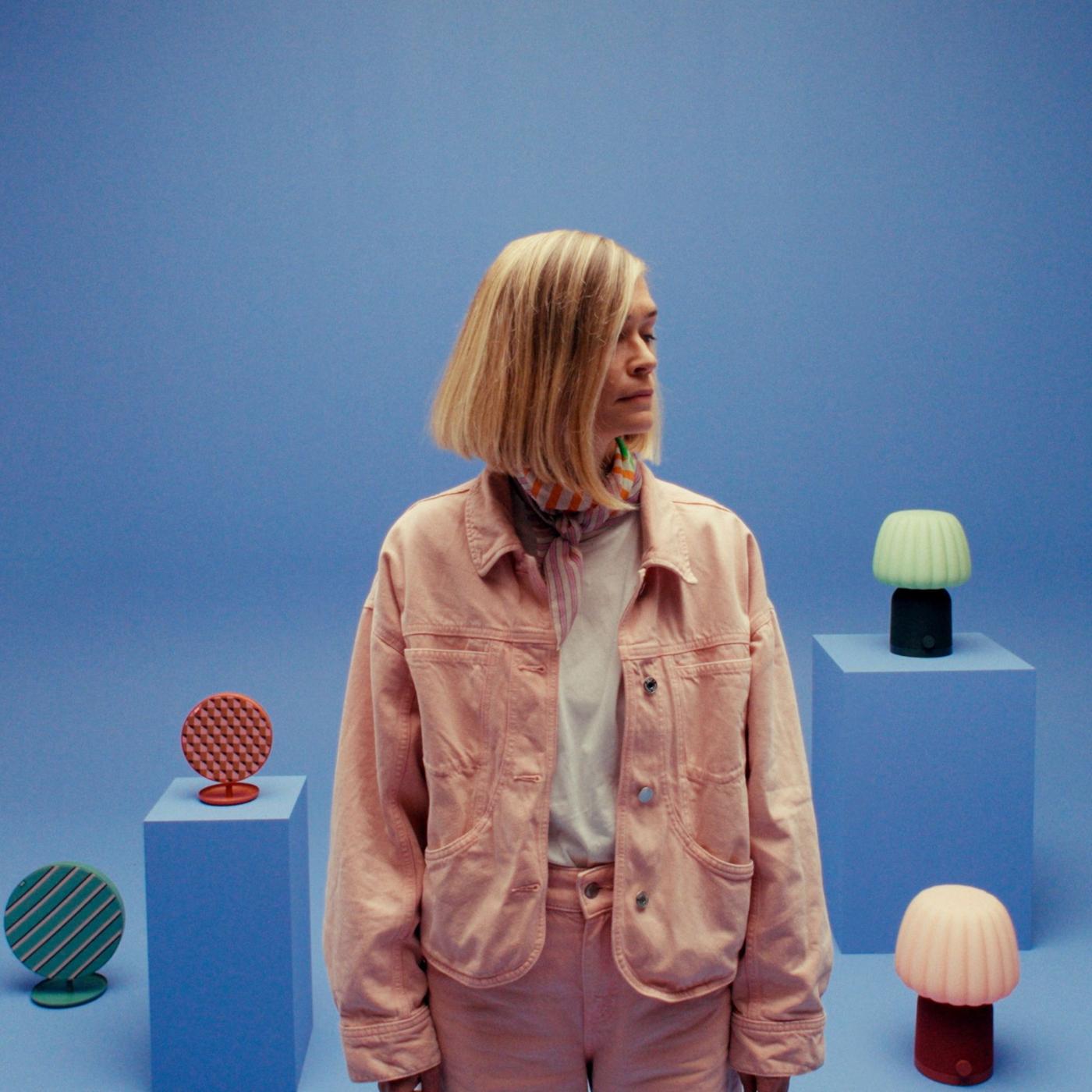 Tekla Severin stands in focus at the front and looking to the side. Two colorful patterned SOLSKYDD speakers and two KULGLASS speakers are displayed on pedestals and the floor behind her.