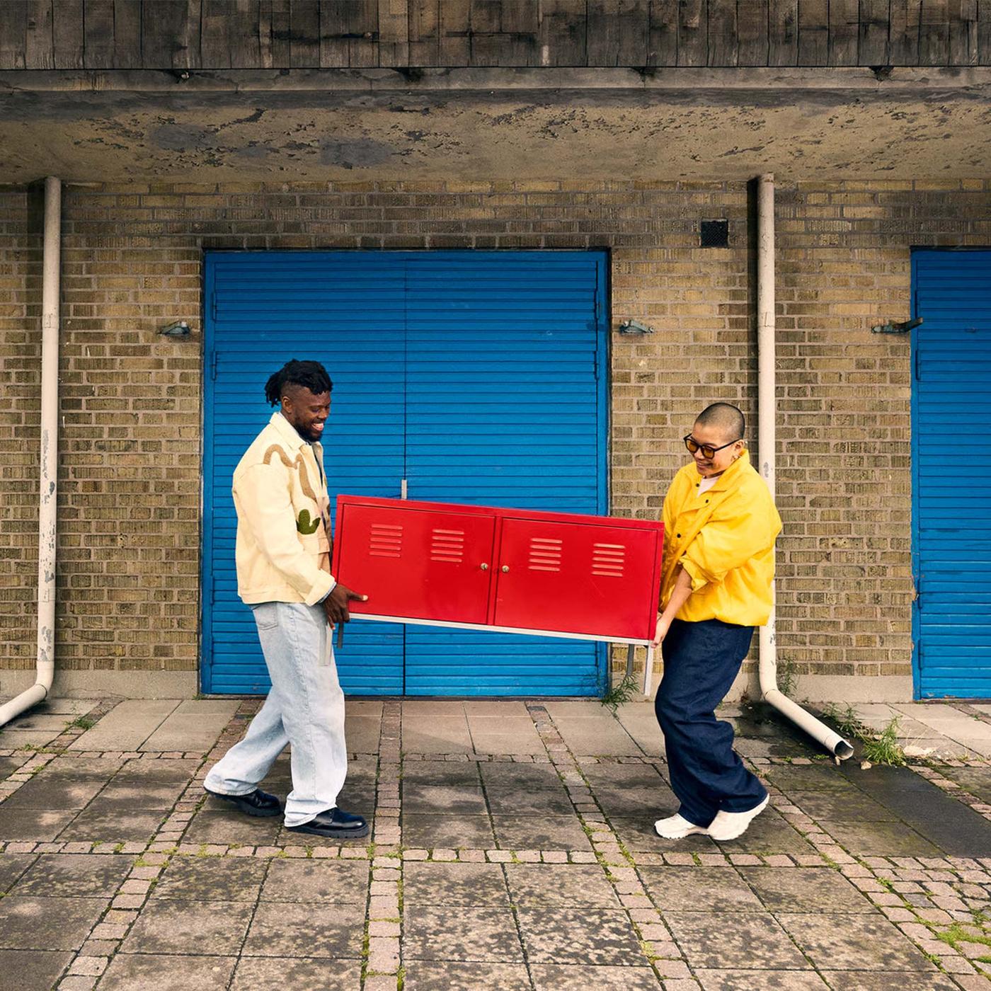 Two people carry a piece of red furniture outside of an apartment building.