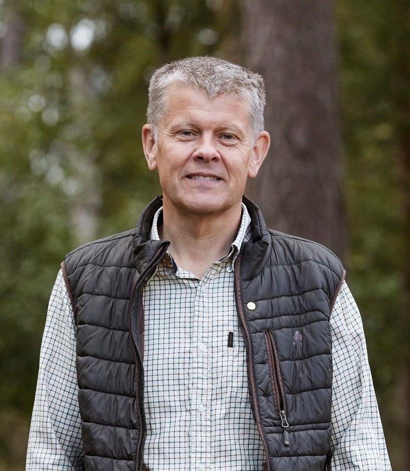 Portrait of Ulf Johansson who wears a checked shirt and a sleeveless puffer jacket, with trees visible in the background.