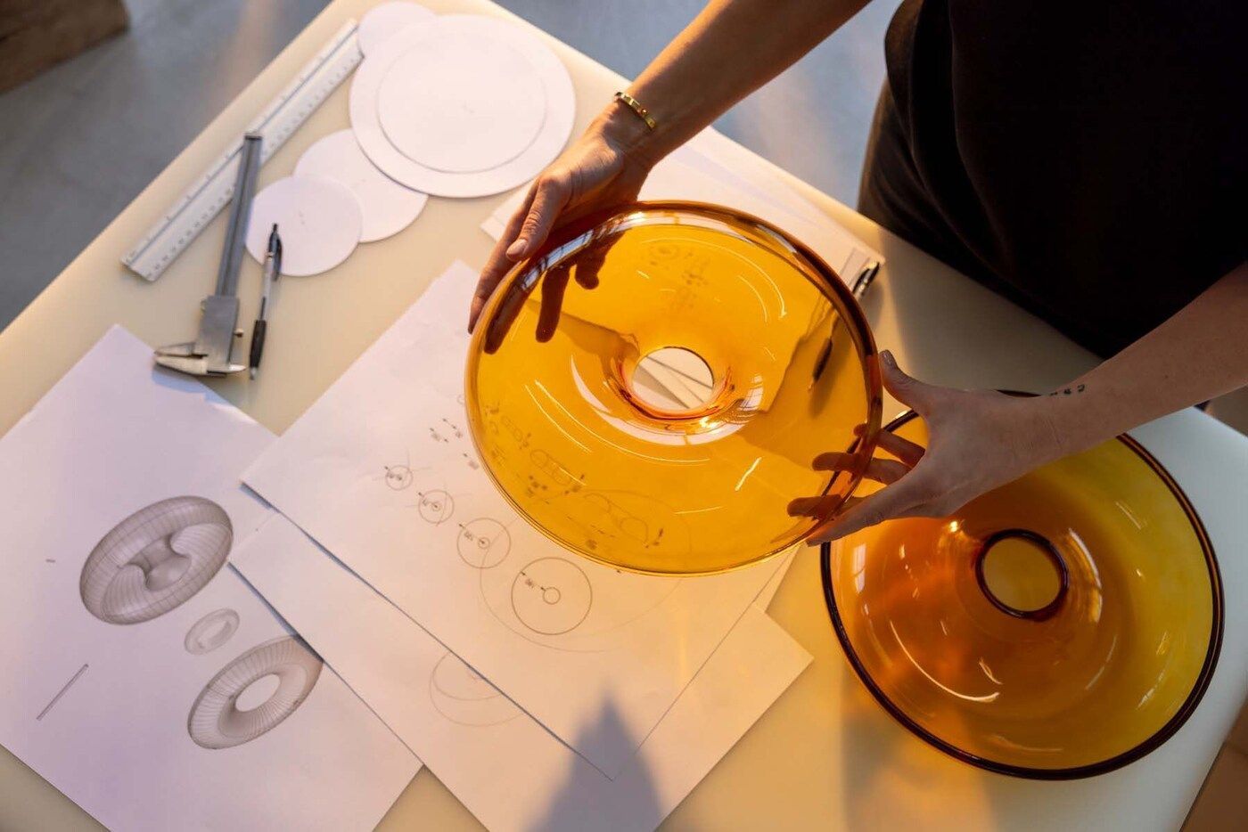 Two doughnut-shaped pieces of orange glassware on a table with design sketches.